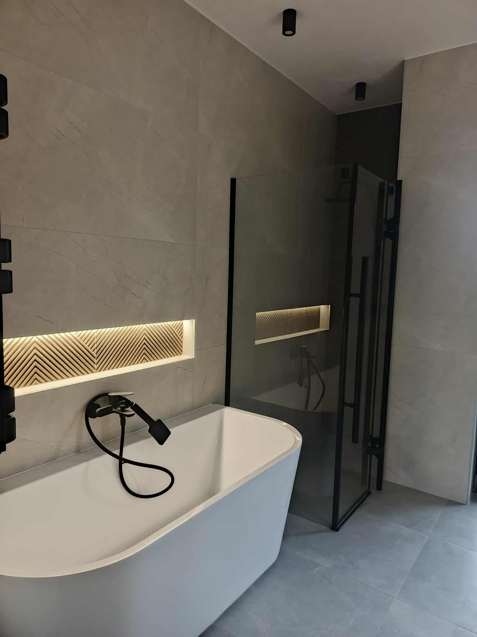 Nowoczesna łazienka z wanną, kabiną prysznicową w czarnej ramie i podświetlaną wnęką z dekoracyjnym wzorem na ścianie z szarych płytek imitujących marmur.