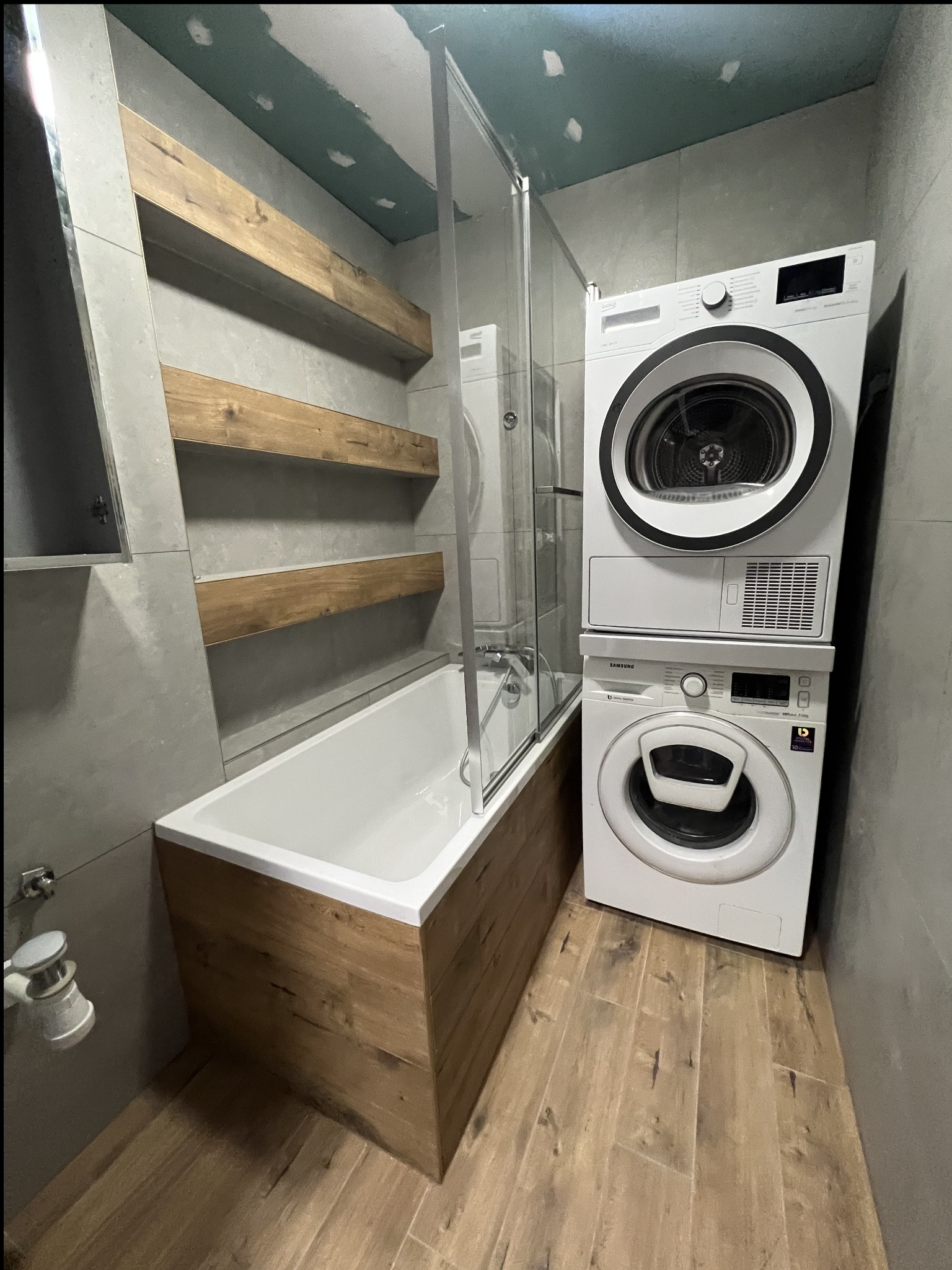 Nowoczesna łazienka po remoncie: wanna z drewnianą obudową, szklany parawan, drewniane półki i pralka z suszarką w szaro-beżowej tonacji.