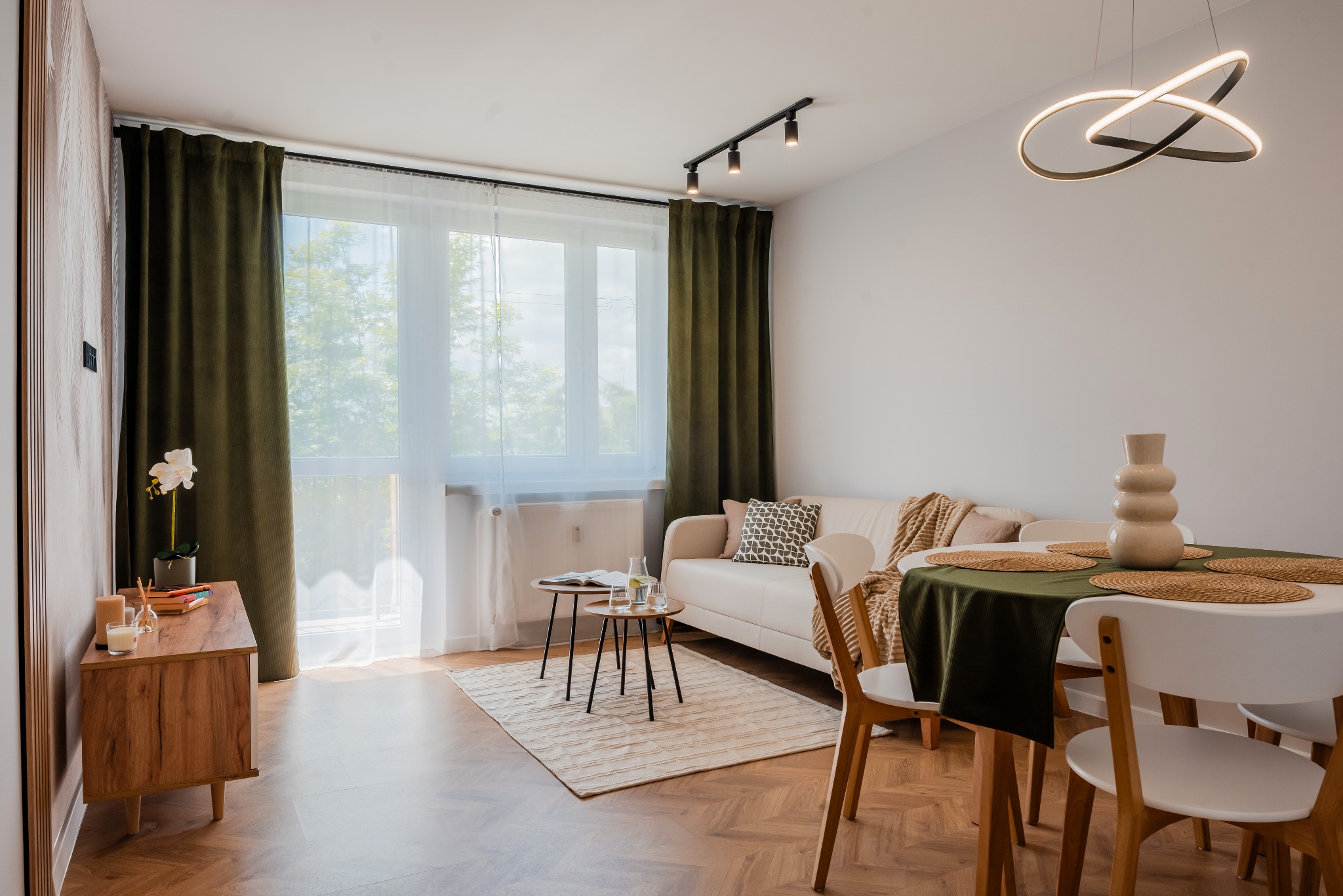 Jasne wnętrze: sofa, stół z zielonym obrusem, drewniana komoda z kwiatami, parkiet w jodełkę, nowoczesna lampa sufitowa i okno z zielonymi zasłonami.