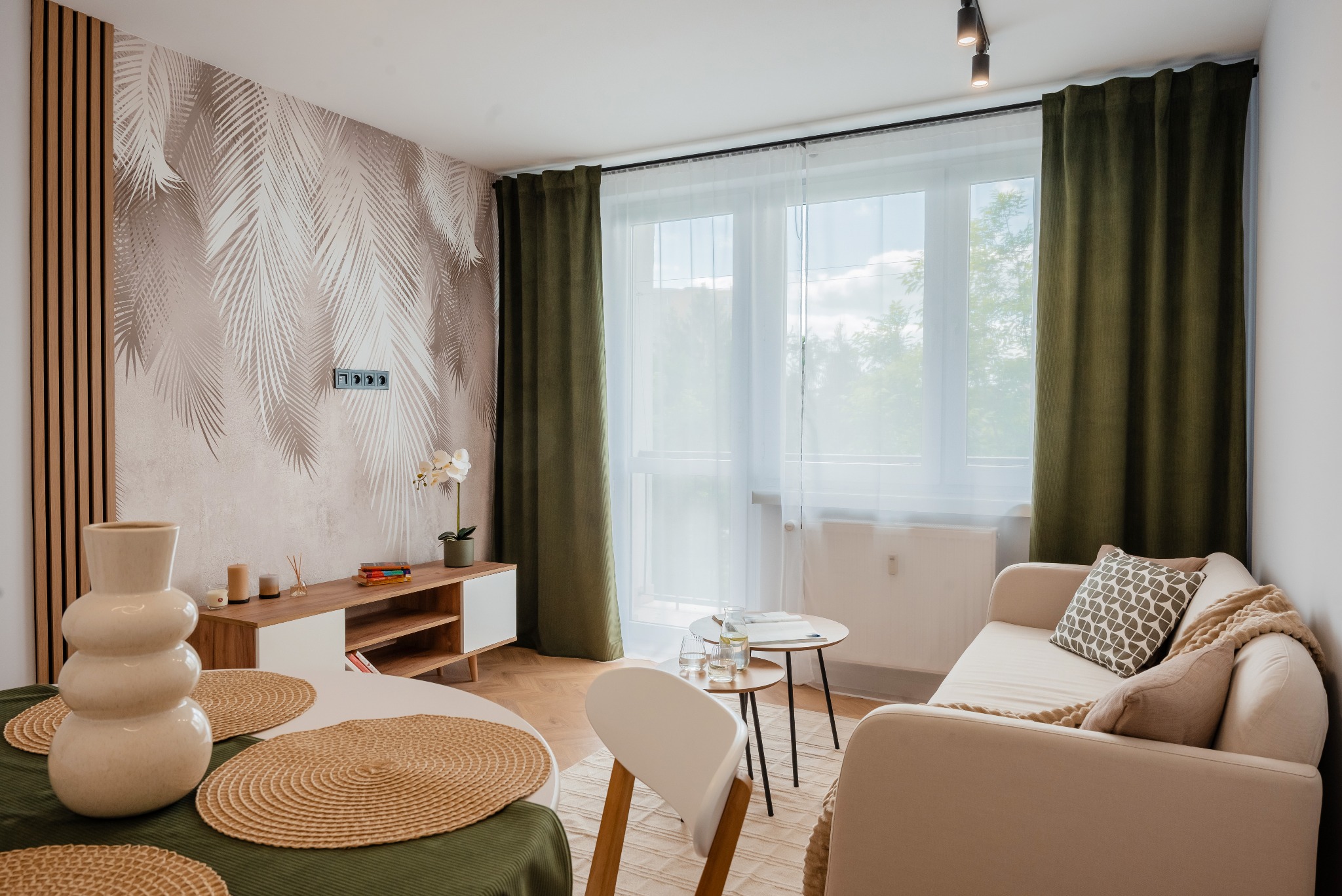 Jasne wnętrze salonu z tapetą w palmy, drewnianą lamelią i beżową sofą. Okno z zielonymi zasłonami i okrągły stół z dekoracjami. Nowoczesny design.