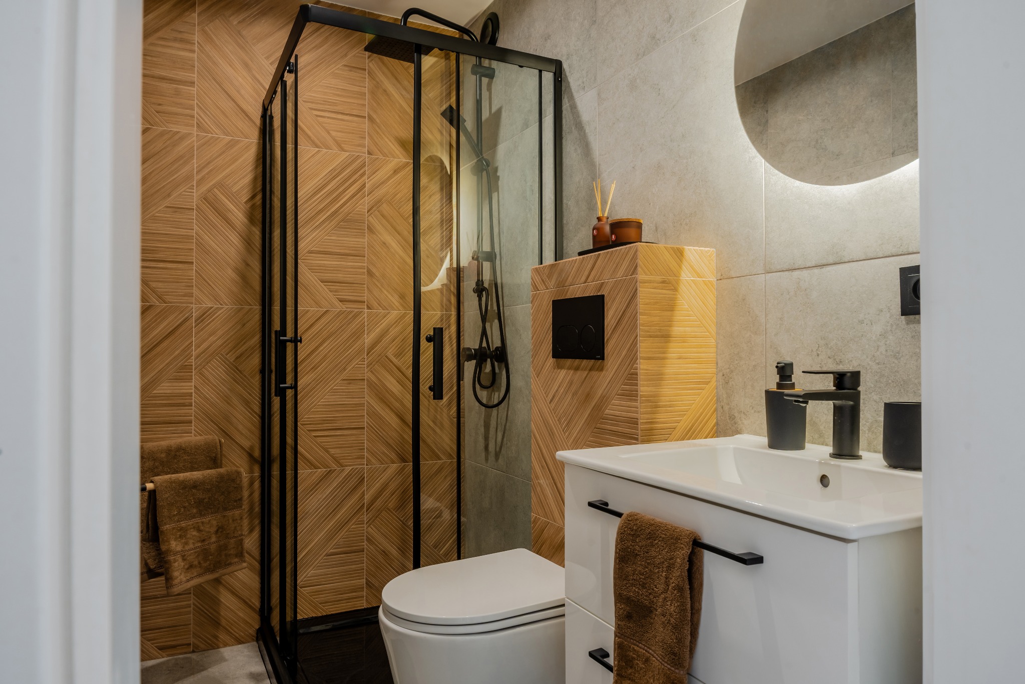 Nowoczesna łazienka z kabiną prysznicową typu walk-in, drewnopodobnymi płytkami i białą szafką umywalkową z czarną armaturą. Okrągłe lustro z podświetleniem.