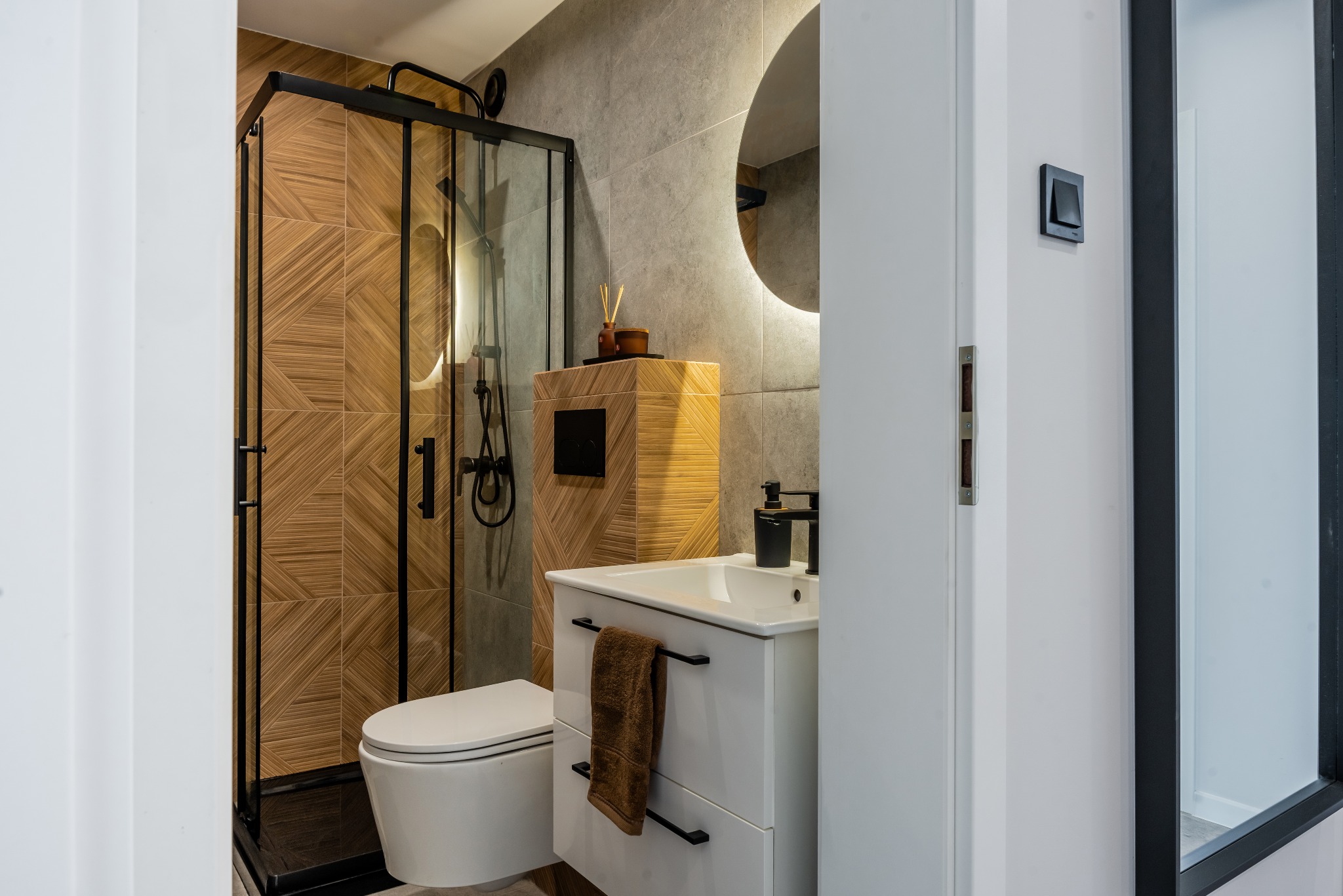 Nowoczesna aranżacja łazienki w Jodłowej: kabina prysznicowa typu walk-in z czarnymi ramami, umywalka z szafką i okrągłe lustro z podświetleniem LED.