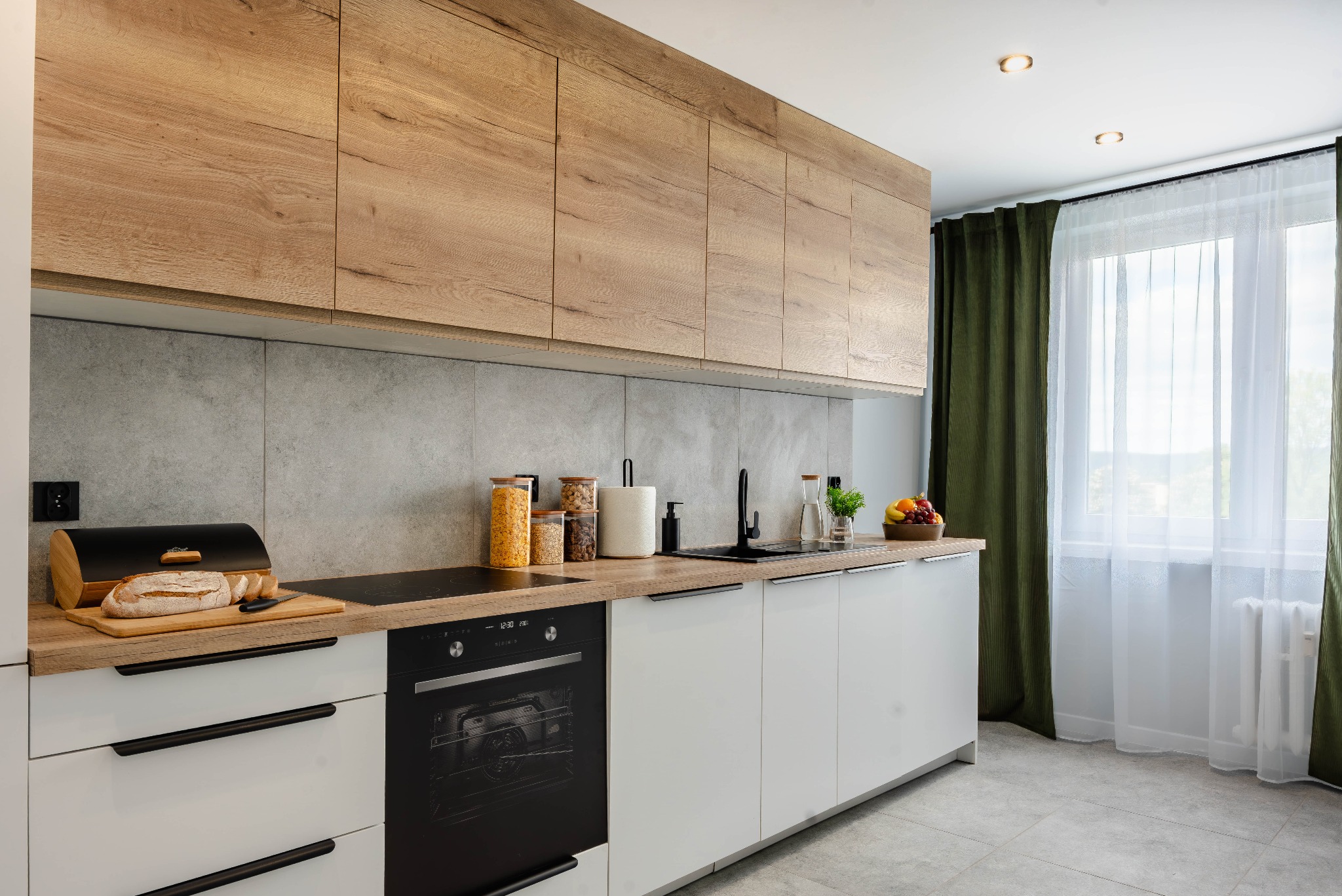 Nowoczesna kuchnia z drewnianymi szafkami górnymi i białymi dolnymi, szarymi płytkami na ścianie i drewnianym blatem. Na blacie chleb, owoce i akcesoria kuchenne.