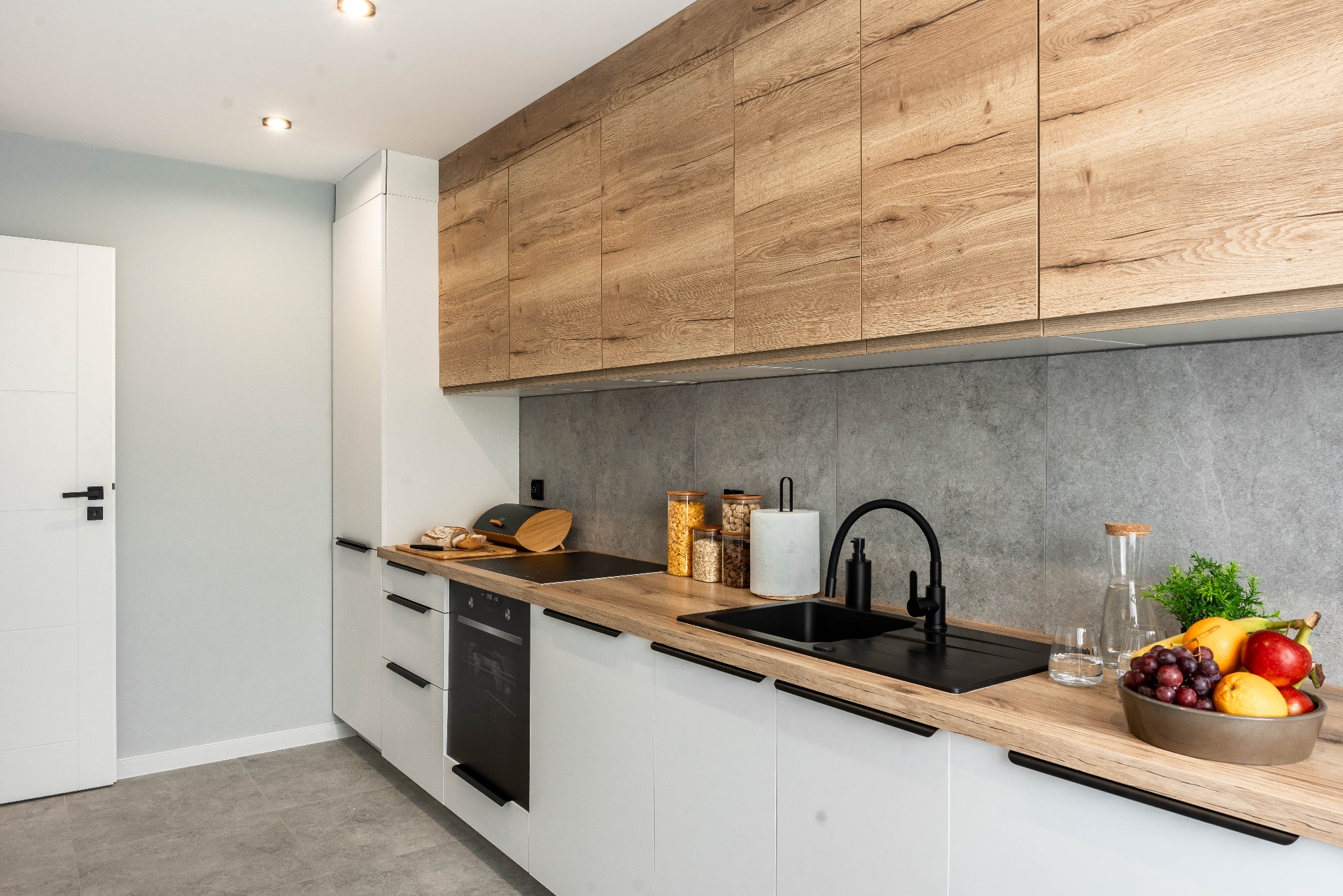 Nowoczesna kuchnia z białymi szafkami dolnymi i drewnopodobnymi górnymi, czarną armaturą i blatem. Na blacie owoce w misie i akcesoria kuchenne.