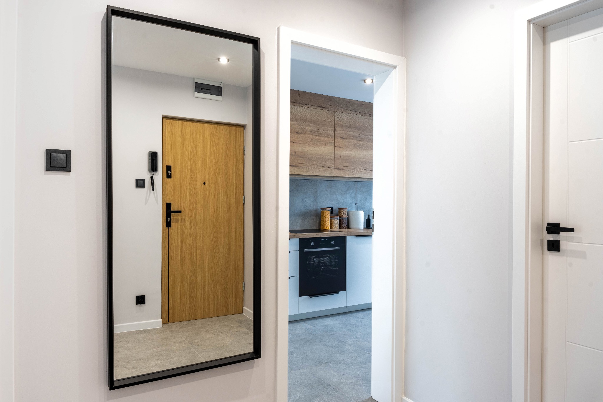 Nowoczesny przedpokój z lustrem w czarnej ramie odbijającym drzwi wejściowe i widokiem na kuchnię. Białe ściany, szara podłoga, minimalistyczny design.