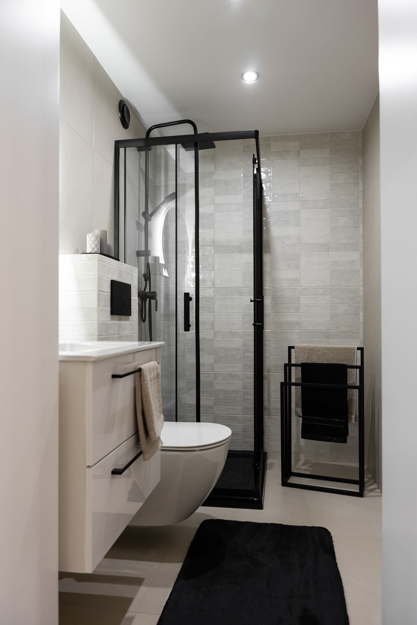 Nowoczesna łazienka z kabiną prysznicową typu walk-in z czarnymi ramami, podwieszaną toaletą i szafką z umywalką. Minimalistyczny design, jasne płytki i czarne akcenty.