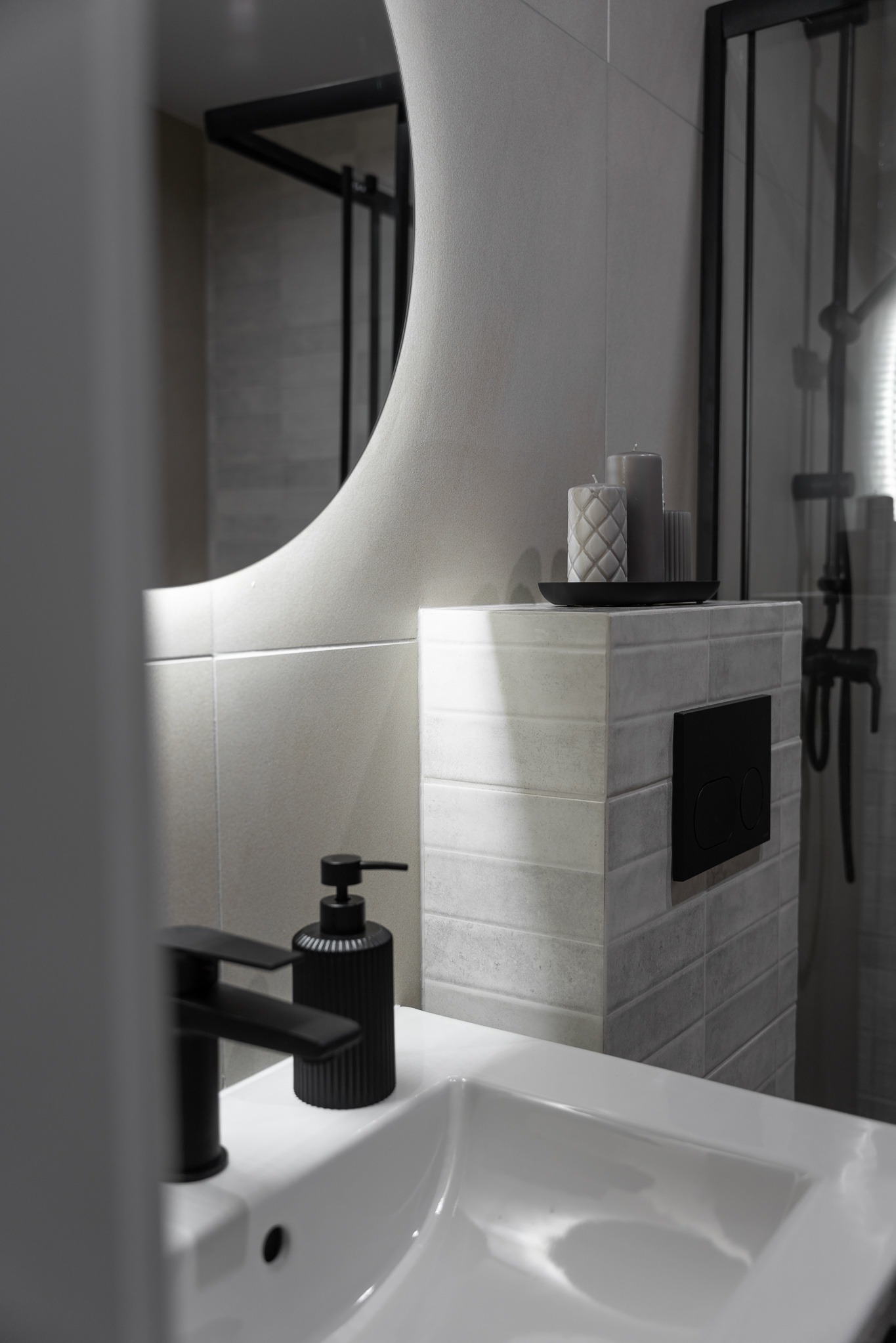 Nowoczesna łazienka: biała umywalka z czarną armaturą i dozownikiem, okrągłe lustro, podwieszana toaleta z płytkami i świecami. Minimalistyczny design.