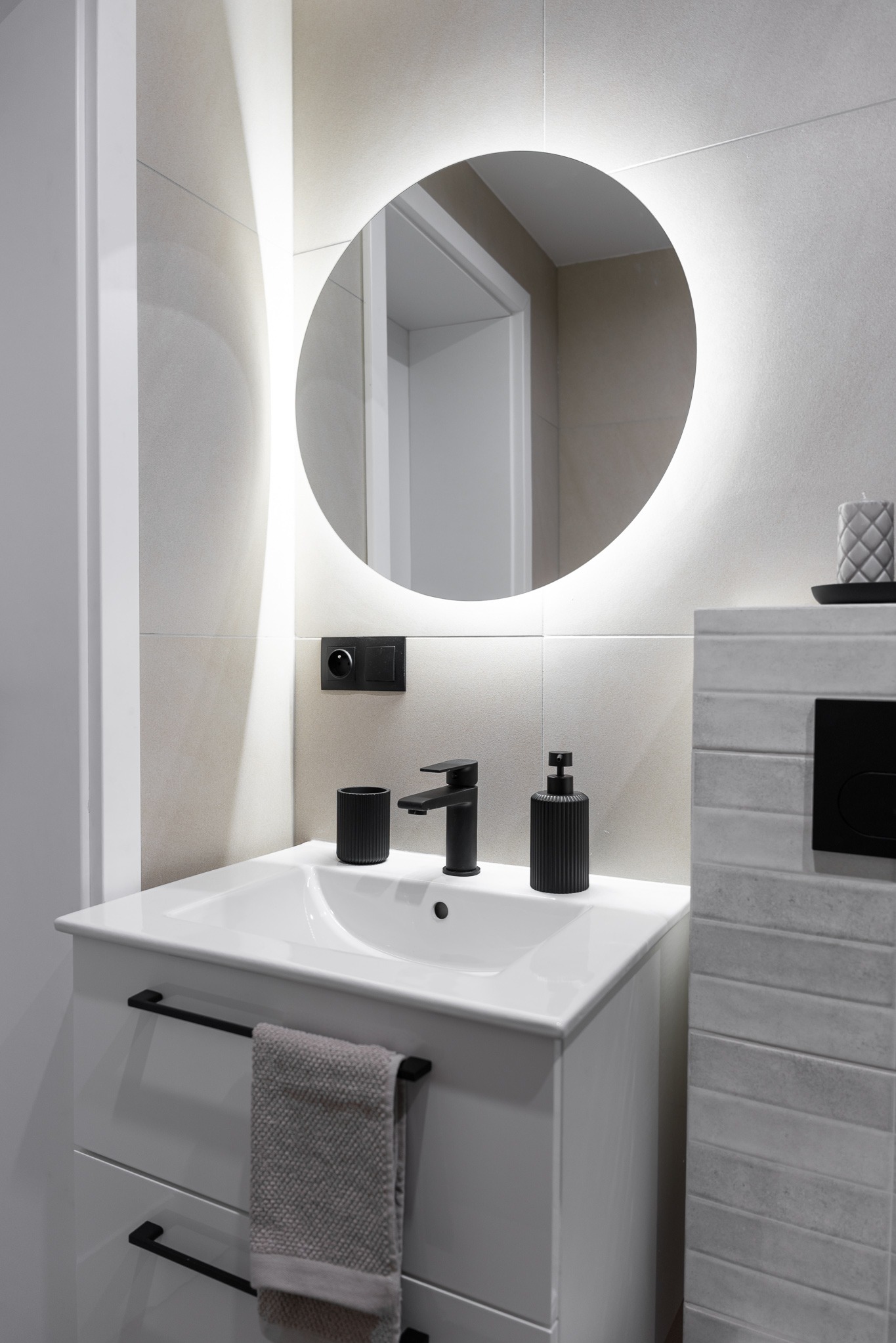 Nowoczesna łazienka: biała szafka z umywalką, czarna bateria i akcesoria, okrągłe lustro z podświetleniem LED, beżowe płytki. Minimalistyczny design.