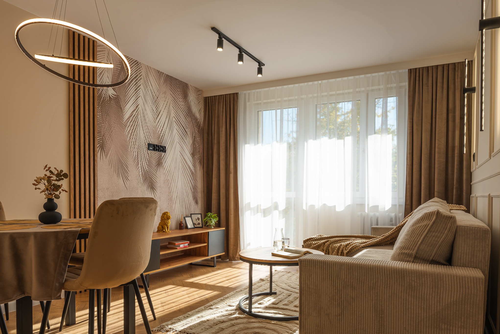 Stylowe wnętrze salonu z jadalnią: beżowa sofa, okrągły stolik, drewniany stół z krzesłami, designerska lampa i ściana z motywem liści palmowych. Nowoczesny design.