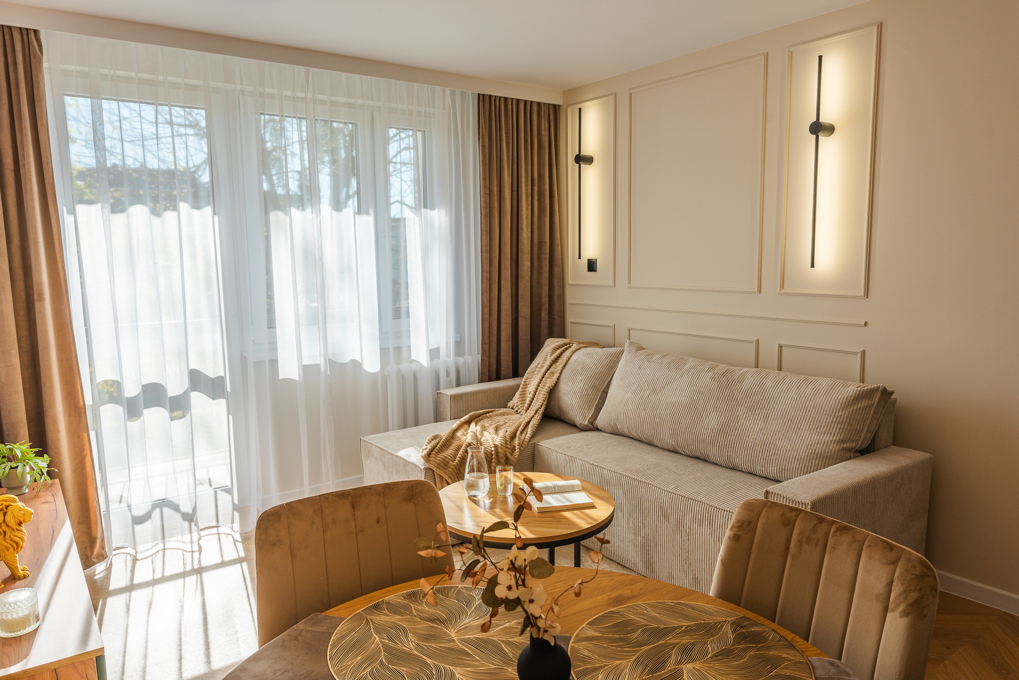 Jasne wnętrze salonu z beżową sofą, okrągłym stolikiem i krzesłami. Na ścianie minimalistyczne, podłużne lampy. Widok na okno z firanami i zasłonami.