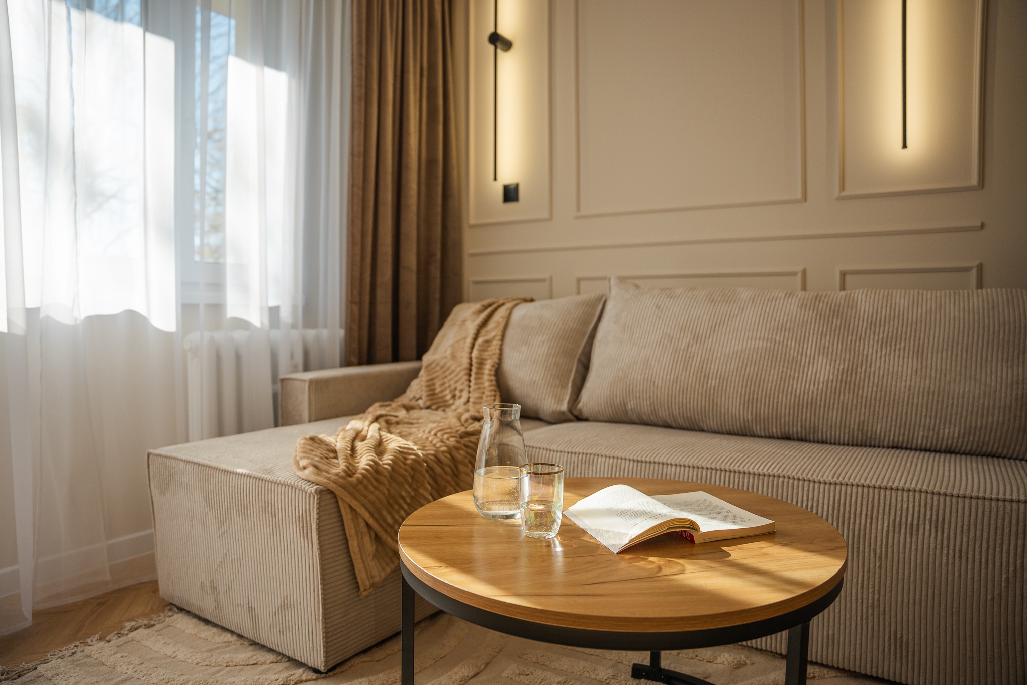 Beżowy narożnik z pluszową narzutą, okrągły stolik z książką i wodą, minimalistyczna lampa ścienna, jasne zasłony w tle. Przytulne, stonowane wnętrze.