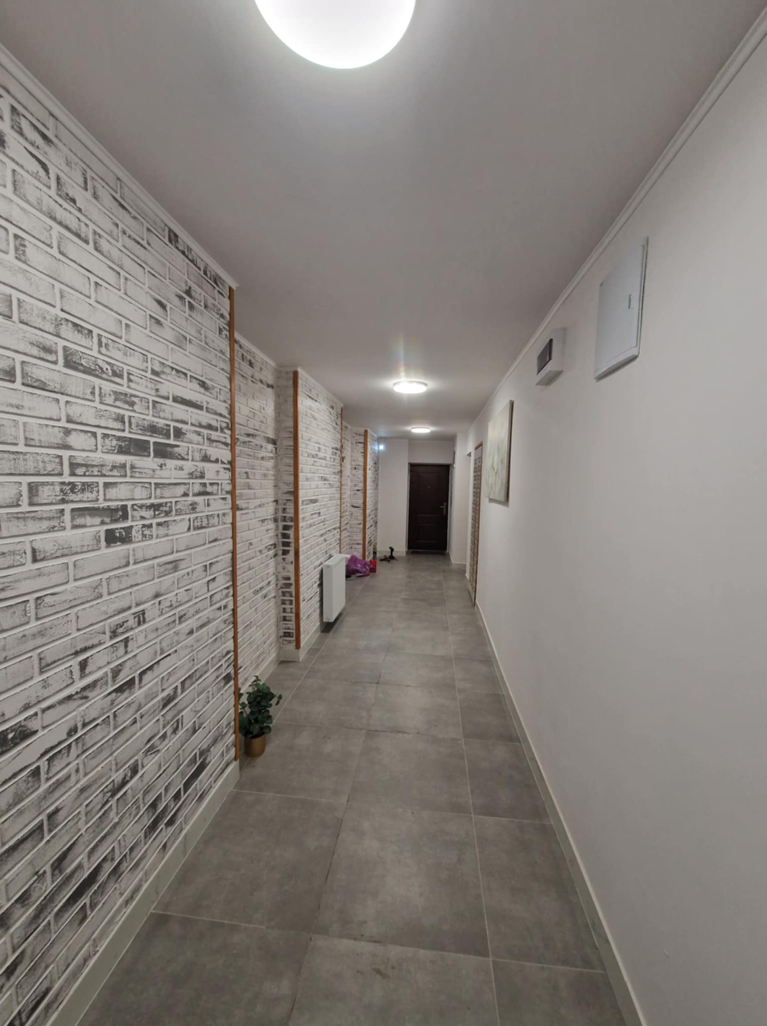 Długi korytarz po remoncie z szarą podłogą i ścianą imitującą białą cegłę. Dekoracyjne listwy pionowe i minimalistyczne oświetlenie sufitowe.