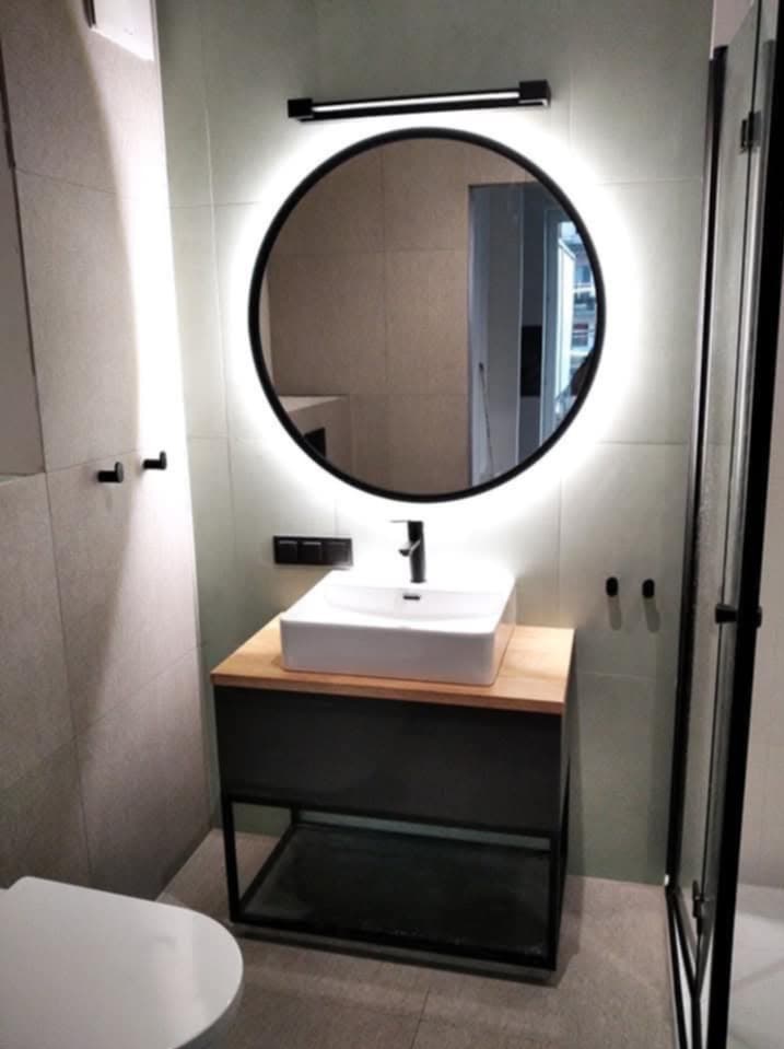 Nowoczesna łazienka z okrągłym lustrem LED, białą umywalką na drewnianym blacie i czarną szafką. Minimalistyczny design, szare płytki i czarne akcenty.