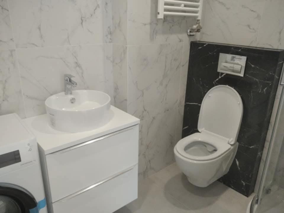 Nowoczesna łazienka po remoncie: biała umywalka nablatowa na szafce, toaleta podwieszana na tle czarnego marmuru, białe płytki imitujące marmur na ścianach.