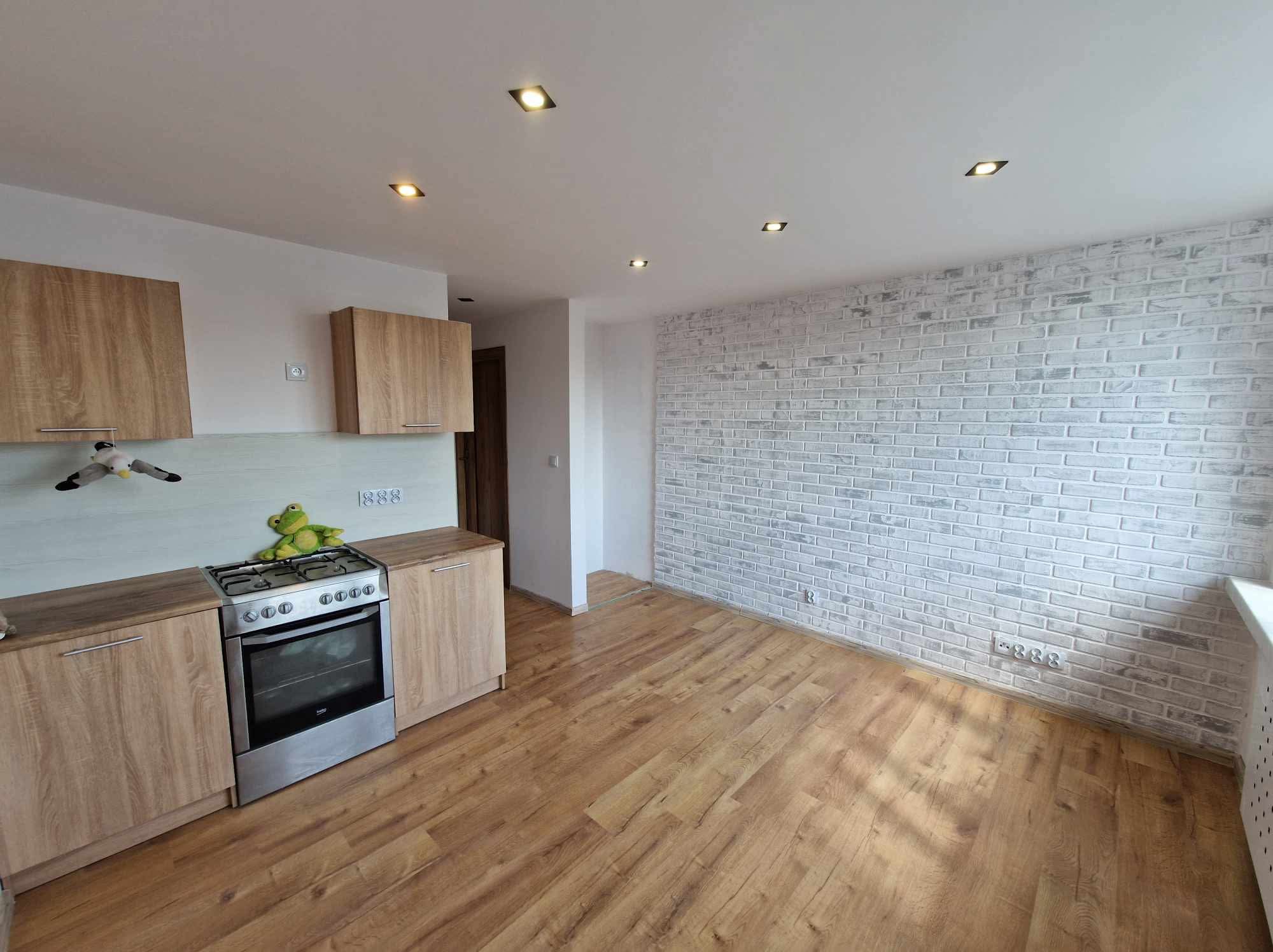 Jasna kuchnia z drewnianymi szafkami i ścianą z białej cegły. Na kuchence pluszowa żaba. Podłoga z paneli imitujących drewno. Nowoczesne oświetlenie sufitowe.