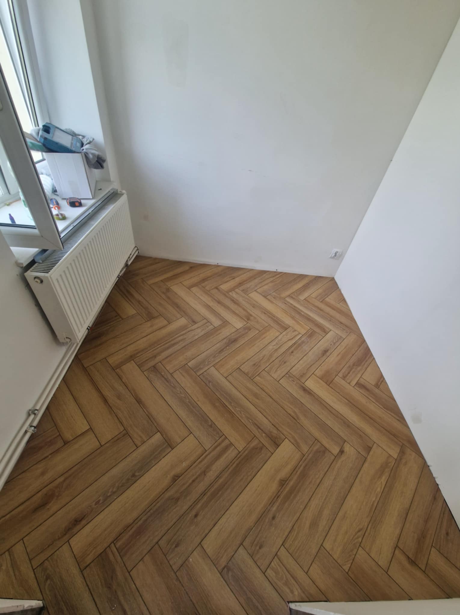 Ukończona podłoga z paneli ułożonych w jodełkę, widok z góry. Białe ściany i grzejnik pod oknem. Jasne, naturalne światło.