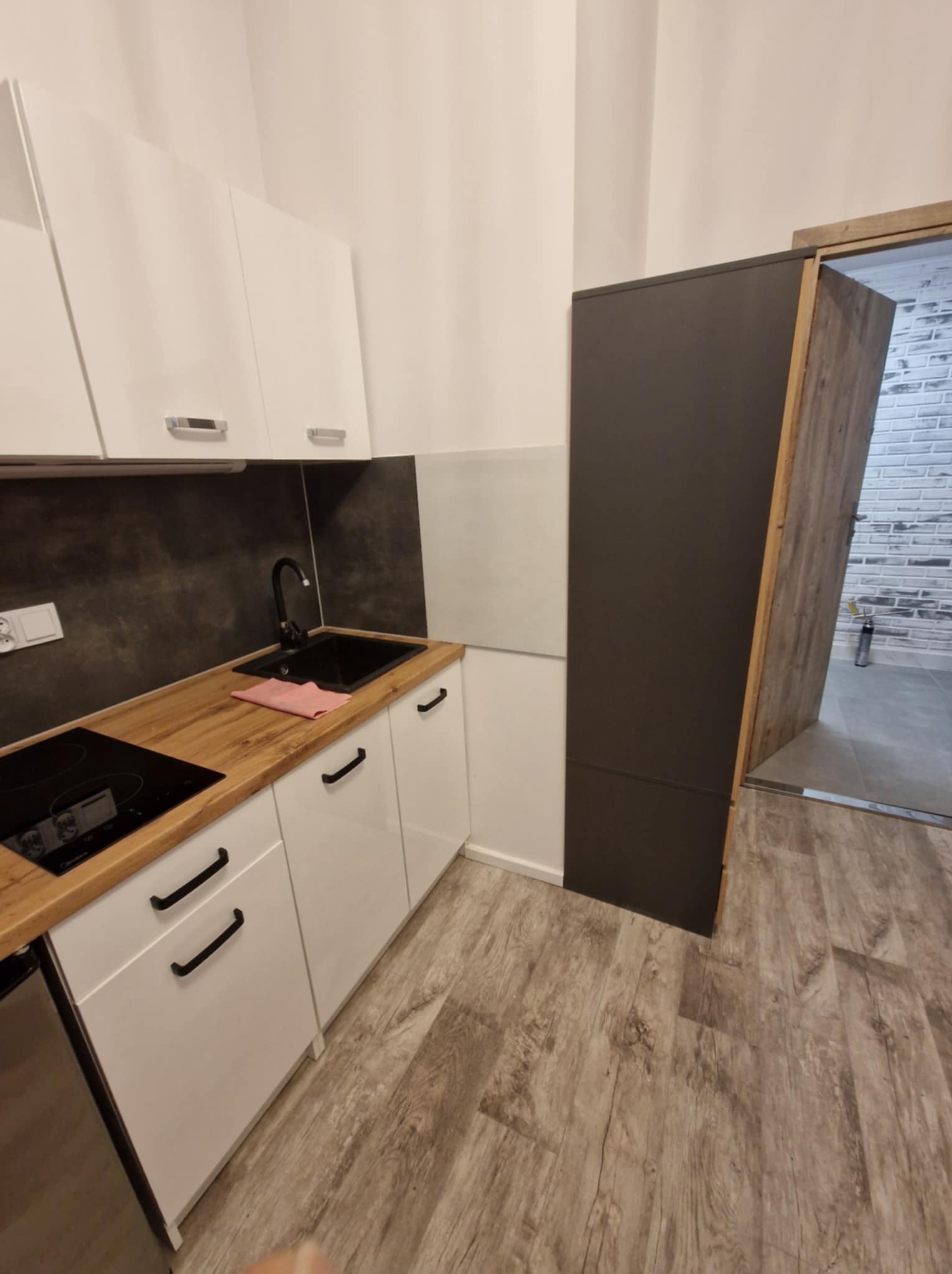 Nowoczesna kuchnia z białymi szafkami, drewnianym blatem i czarnymi akcentami. Widoczny zlew, płyta indukcyjna i fragment otwartych drzwi do innego pomieszczenia.