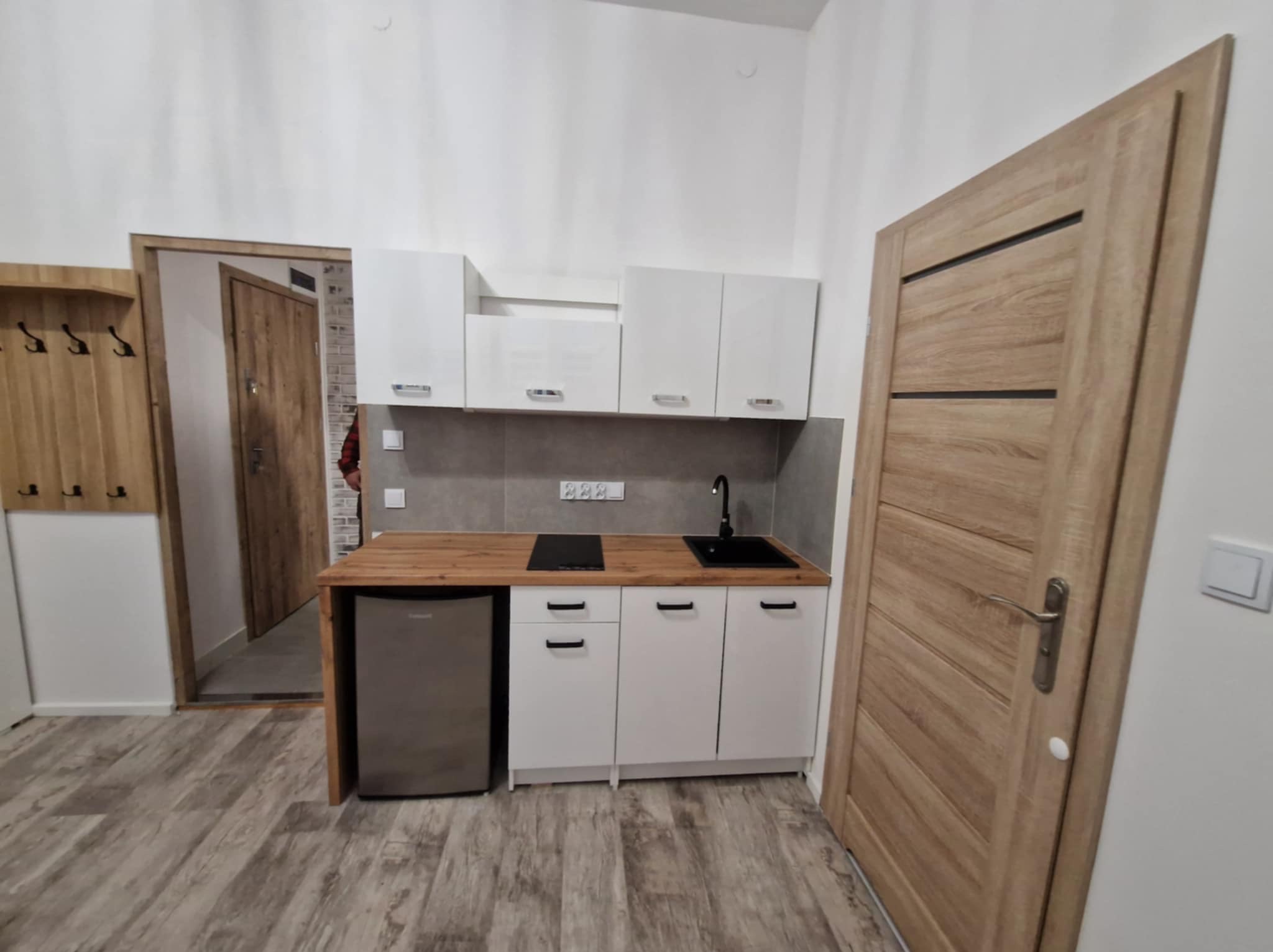 Nowoczesna, mała kuchnia z białymi szafkami, drewnianym blatem i małą lodówką. Widoczna drewniana podłoga i drzwi z drewnianą okleiną. Minimalistyczny design.