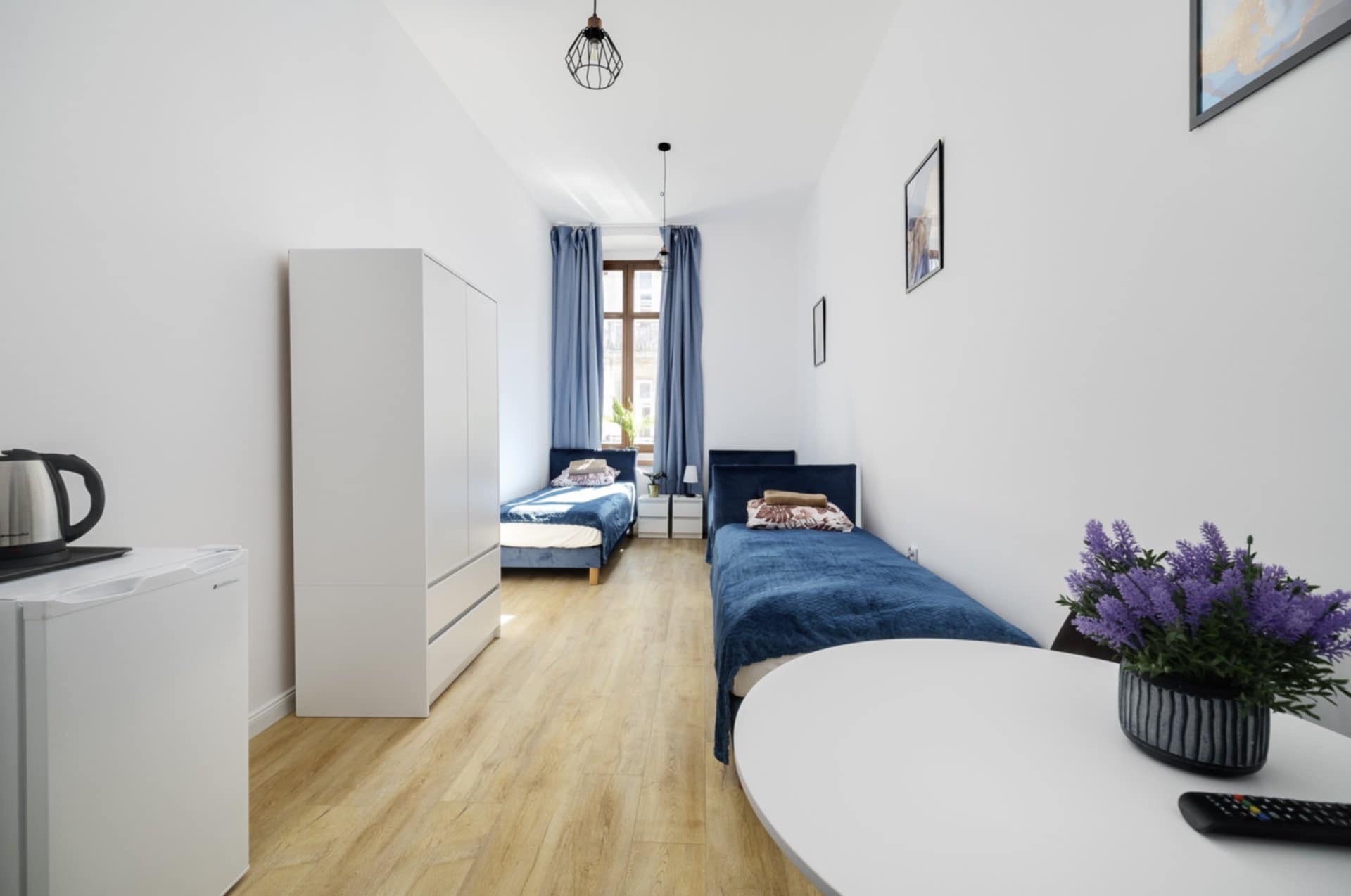 Jasne wnętrze pokoju z dwoma łóżkami, szafą i stolikiem z kwiatami. Minimalistyczny design, drewniana podłoga, niebieskie zasłony i pościel. Widok na okno.