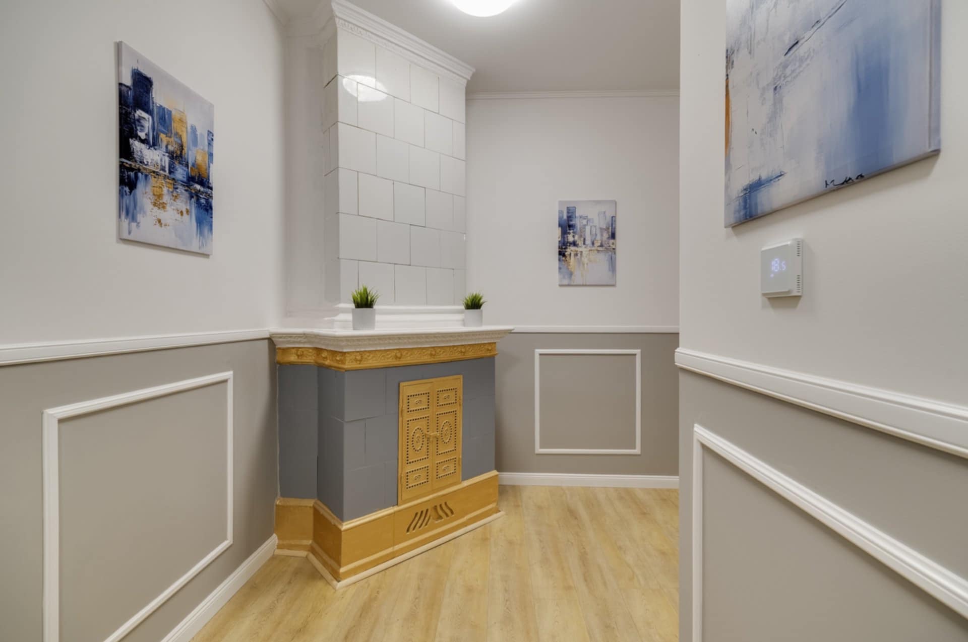 Wykończone wnętrze z ozdobnym piecem kaflowym, sztukaterią na ścianach i drewnianą podłogą. Na ścianach wiszą obrazy, a na piecu stoją małe rośliny.