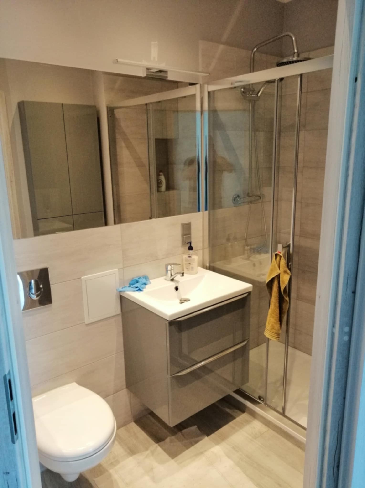 Nowoczesna łazienka po renowacji: szara szafka z umywalką, biała toaleta, kabina prysznicowa z żółtym ręcznikiem i lustro z szafką w tle. Jasne płytki na ścianach.