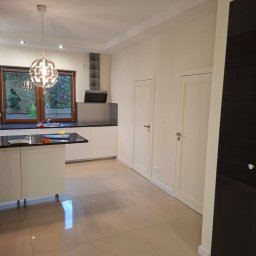 GryCars Michał Grykałowski - Jasna kuchnia po remoncie: beżowa podłoga, białe ściany i meble, czarny blat. Okno z widokiem na zieleń. Nowoczesna lampa wisząca.