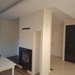 GryCars Michał Grykałowski - Jasne wnętrze po remoncie: kremowe ściany, czarny wkład kominkowy z szybą, biały stół z nogami, beżowa podłoga. Widoczne listwy przypodłogowe i drzwi.