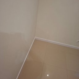 GryCars Michał Grykałowski - Beżowe płytki podłogowe w rogu pokoju, ściany pomalowane na jasny odcień beżu, biała listwa przypodłogowa. Widok z góry, neutralne oświetlenie.