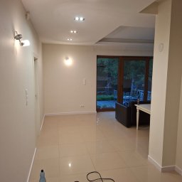 GryCars Michał Grykałowski - Jasne, przestronne wnętrze po remoncie z beżowymi płytkami podłogowymi, nowoczesnym oświetleniem sufitowym i widokiem na ogród przez przeszklone drzwi.