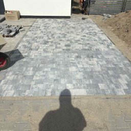 GryCars Michał Grykałowski - Ułożona szara kostka brukowa na podjeździe domu, widoczni pracownicy w oddali i sterta kostki obok budynku. Cień osoby w dolnej części kadru.