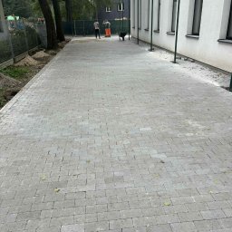 GryCars Michał Grykałowski - Ułożona szara kostka brukowa na długim chodniku przy budynku. W tle pracownicy i taczka. Widoczne opadłe liście na kostce.