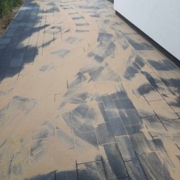 GryCars Michał Grykałowski - Ciemnoszara kostka brukowa zasypana piaskiem kwarcowym, przygotowana do zagęszczenia. Widoczny fragment budynku z białą elewacją i osoba z miotłą w tle.