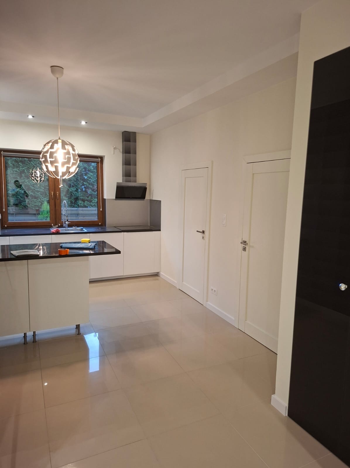 Jasna kuchnia po remoncie: beżowa podłoga, białe ściany i meble, czarny blat. Okno z widokiem na zieleń. Nowoczesna lampa wisząca.