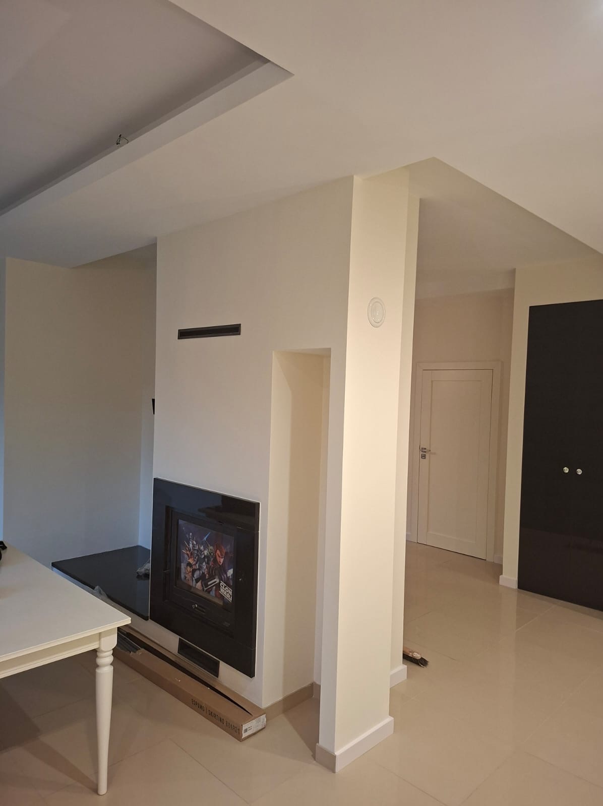 Jasne wnętrze po remoncie: kremowe ściany, czarny wkład kominkowy z szybą, biały stół z nogami, beżowa podłoga. Widoczne listwy przypodłogowe i drzwi.