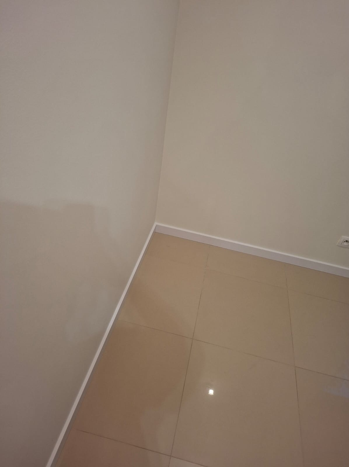 Beżowe płytki podłogowe w rogu pokoju, ściany pomalowane na jasny odcień beżu, biała listwa przypodłogowa. Widok z góry, neutralne oświetlenie.