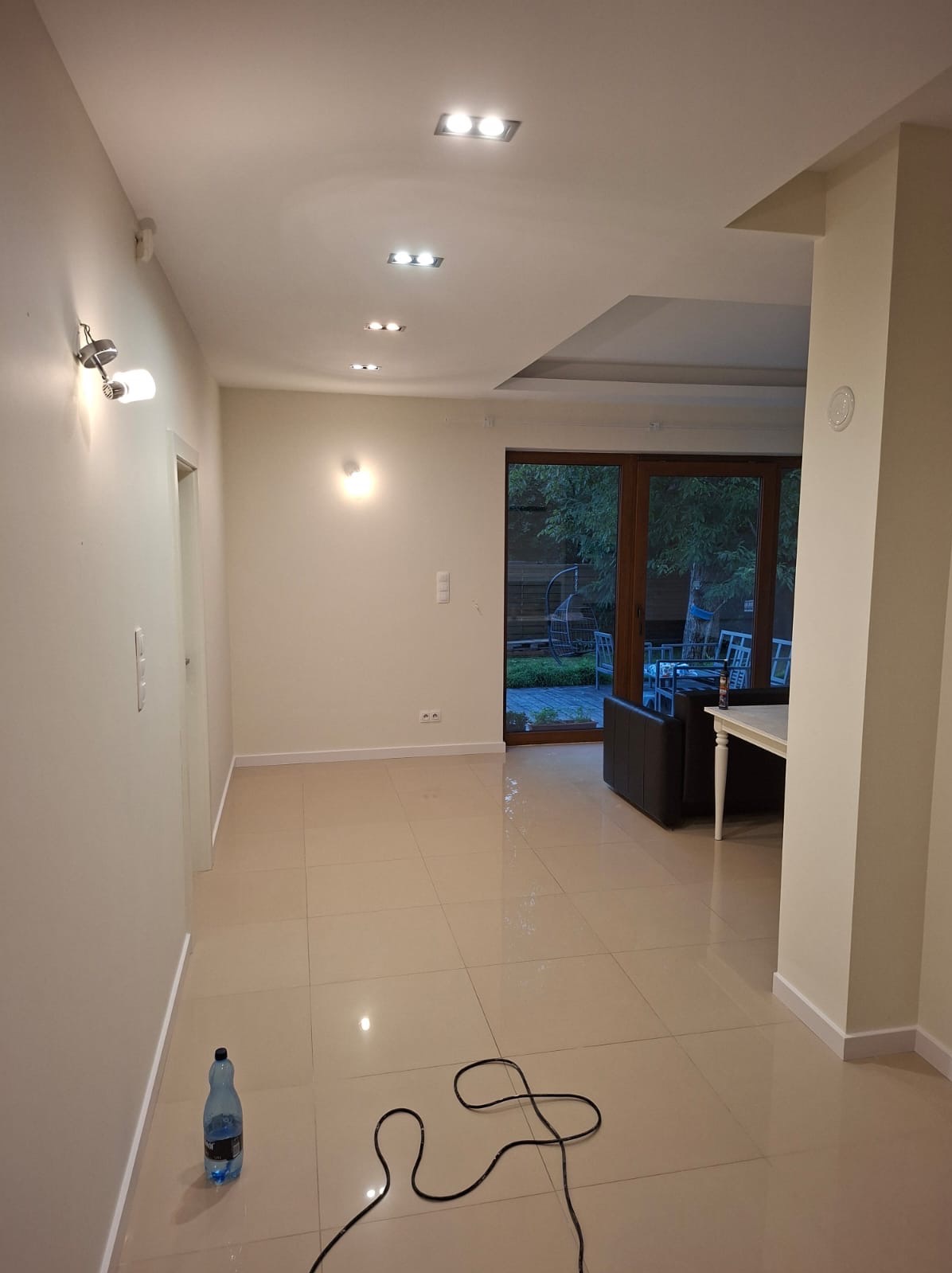 Jasne, przestronne wnętrze po remoncie z beżowymi płytkami podłogowymi, nowoczesnym oświetleniem sufitowym i widokiem na ogród przez przeszklone drzwi.