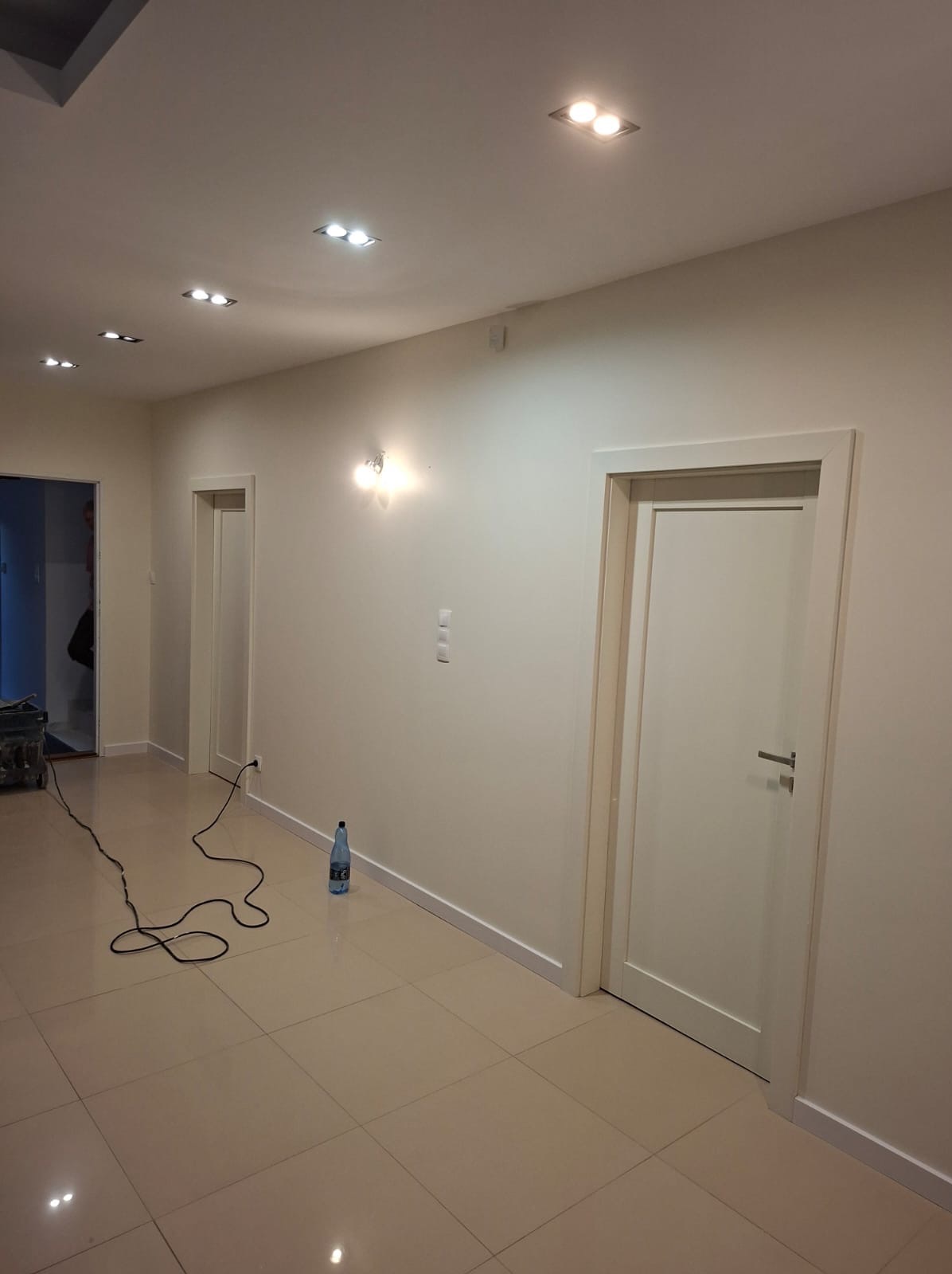 Jasne wnętrze po remoncie: świeżo pomalowane ściany, nowe drzwi z srebrną klamką, błyszcząca podłoga z płytek. Kabel od sprzętu malarskiego na podłodze.