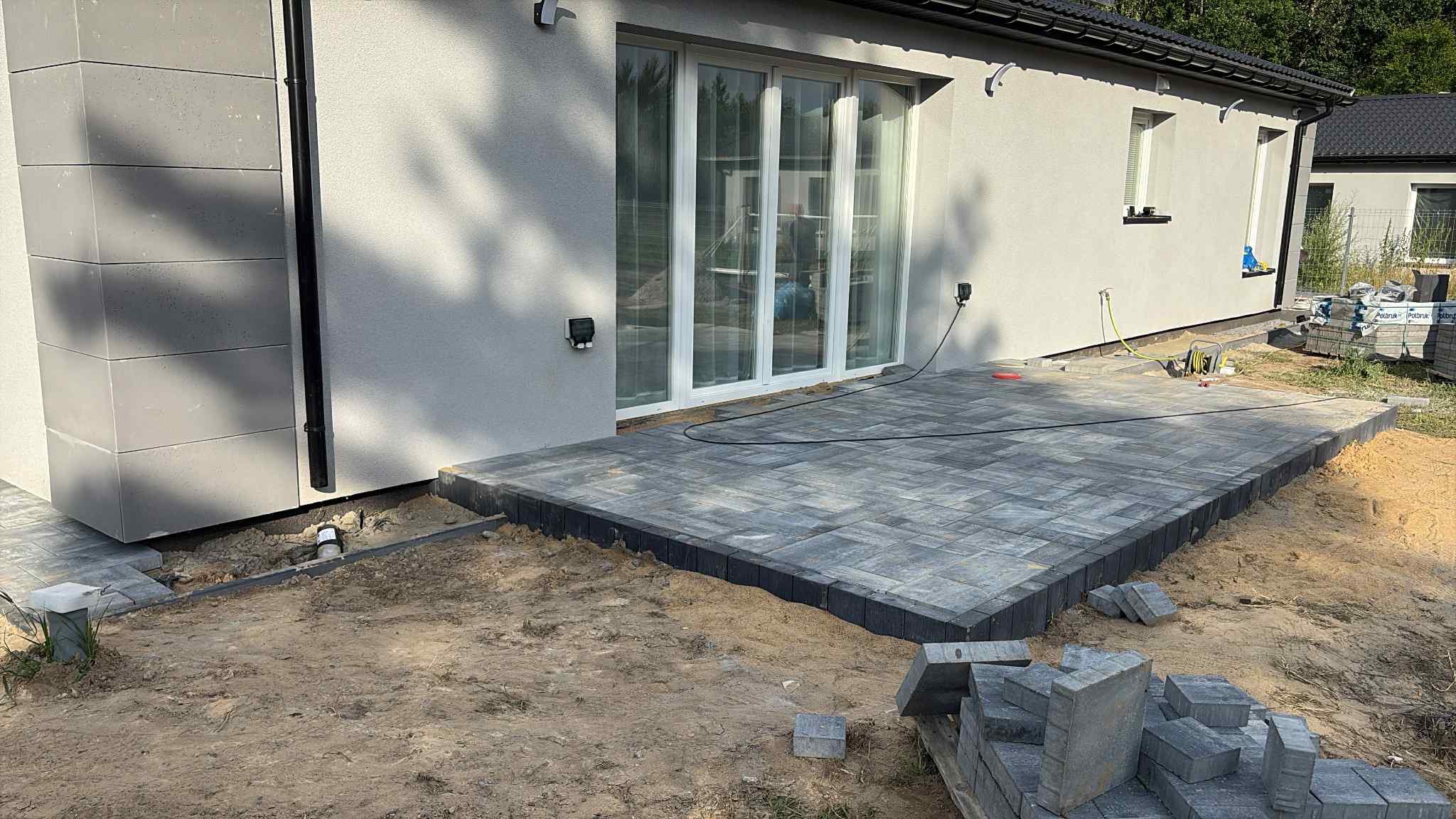Szara kostka brukowa ułożona na tarasie domu jednorodzinnego. Widoczne palety z kostką, przygotowane podłoże i fragment elewacji z oknami. Ujęcie z poziomu gruntu.