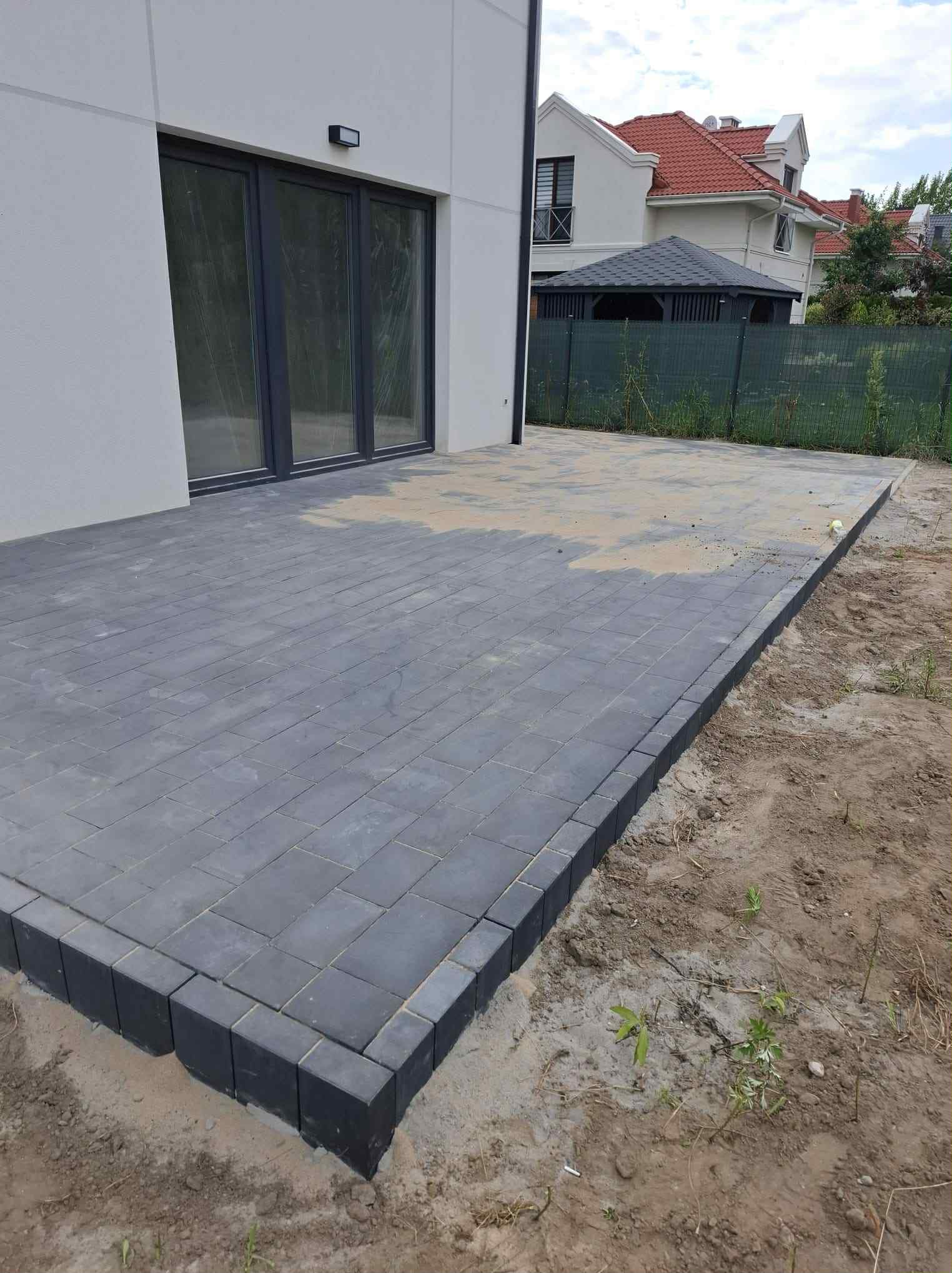 Nowoczesny taras z szarej kostki brukowej, obrzeża z ciemnych kostek. Widok na dom z dużymi oknami i zielony płot w tle. Minimalistyczny design.