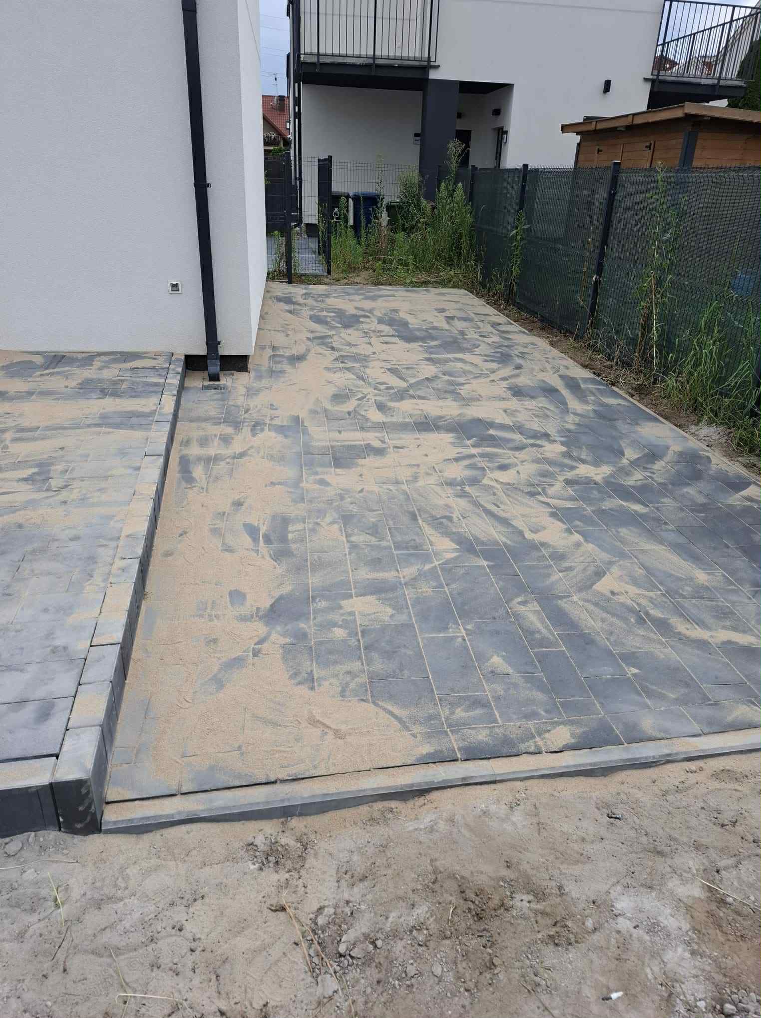 Ułożona, szara kostka brukowa na posesji, zasypana piaskiem. Widoczne obrzeża i fragment budynku z rynną. Tło: ogrodzenie z siatki i zieleń.