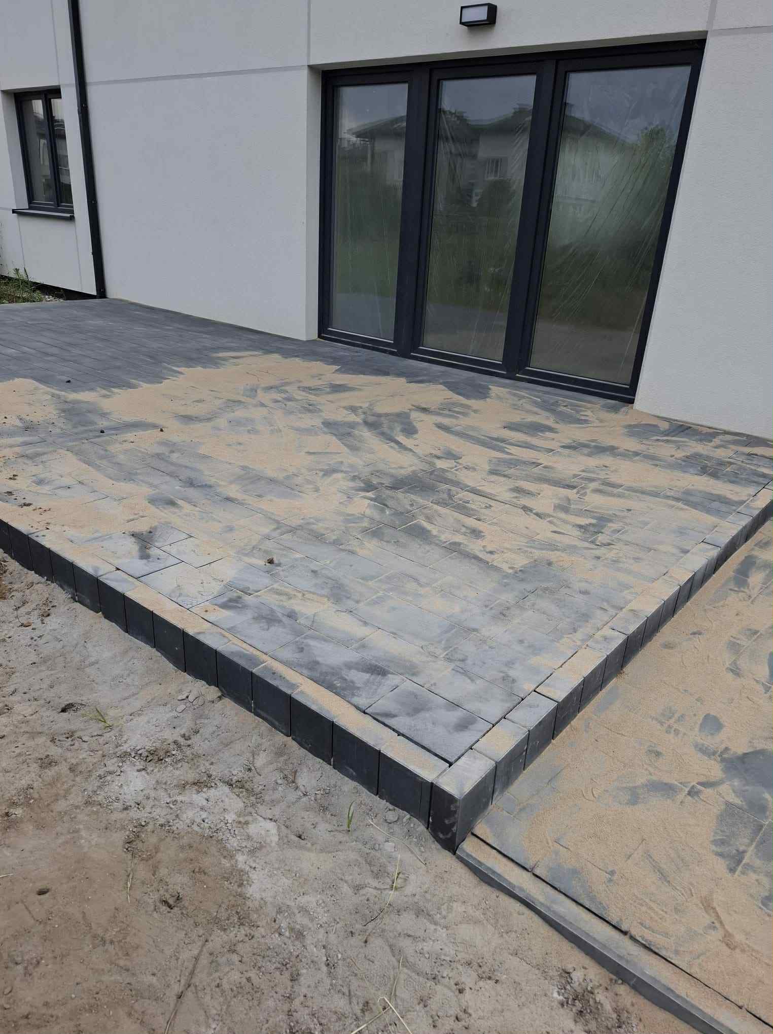 Ułożona, szara kostka brukowa na tarasie w Markach, z widocznymi śladami piasku i obrzeżami. Nowoczesny dom z dużymi oknami w tle. Widok z poziomu gruntu.