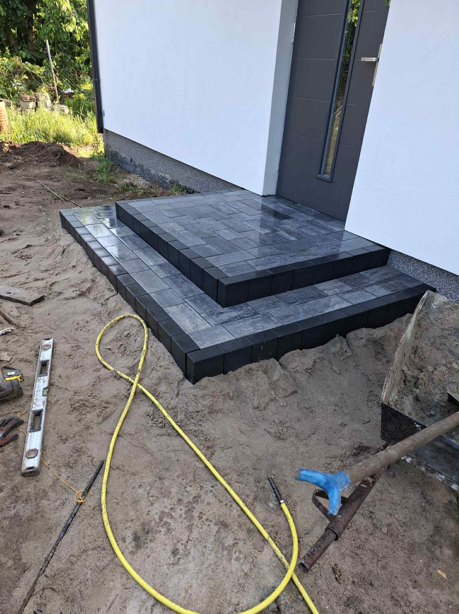 Schody wejściowe z ciemnej kostki brukowej, częściowo ułożone na piasku. Widoczne narzędzia brukarskie: poziomica, łopata i wąż. Tło stanowi biała ściana budynku z szarymi drzwiami.