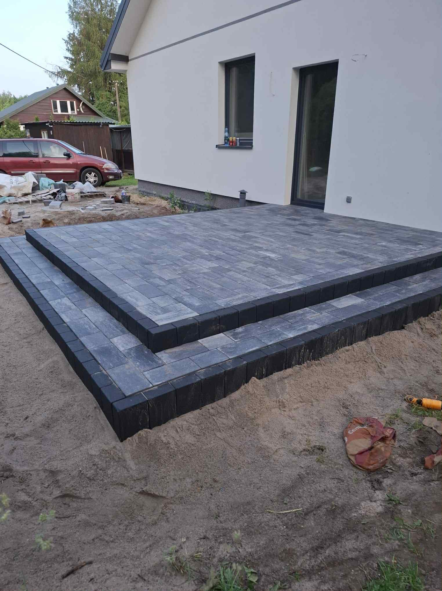 Nowy taras z szarej kostki brukowej z czarnym obramowaniem, prowadzący do białego domu. Widoczne worki z materiałami budowlanymi i narzędzia.