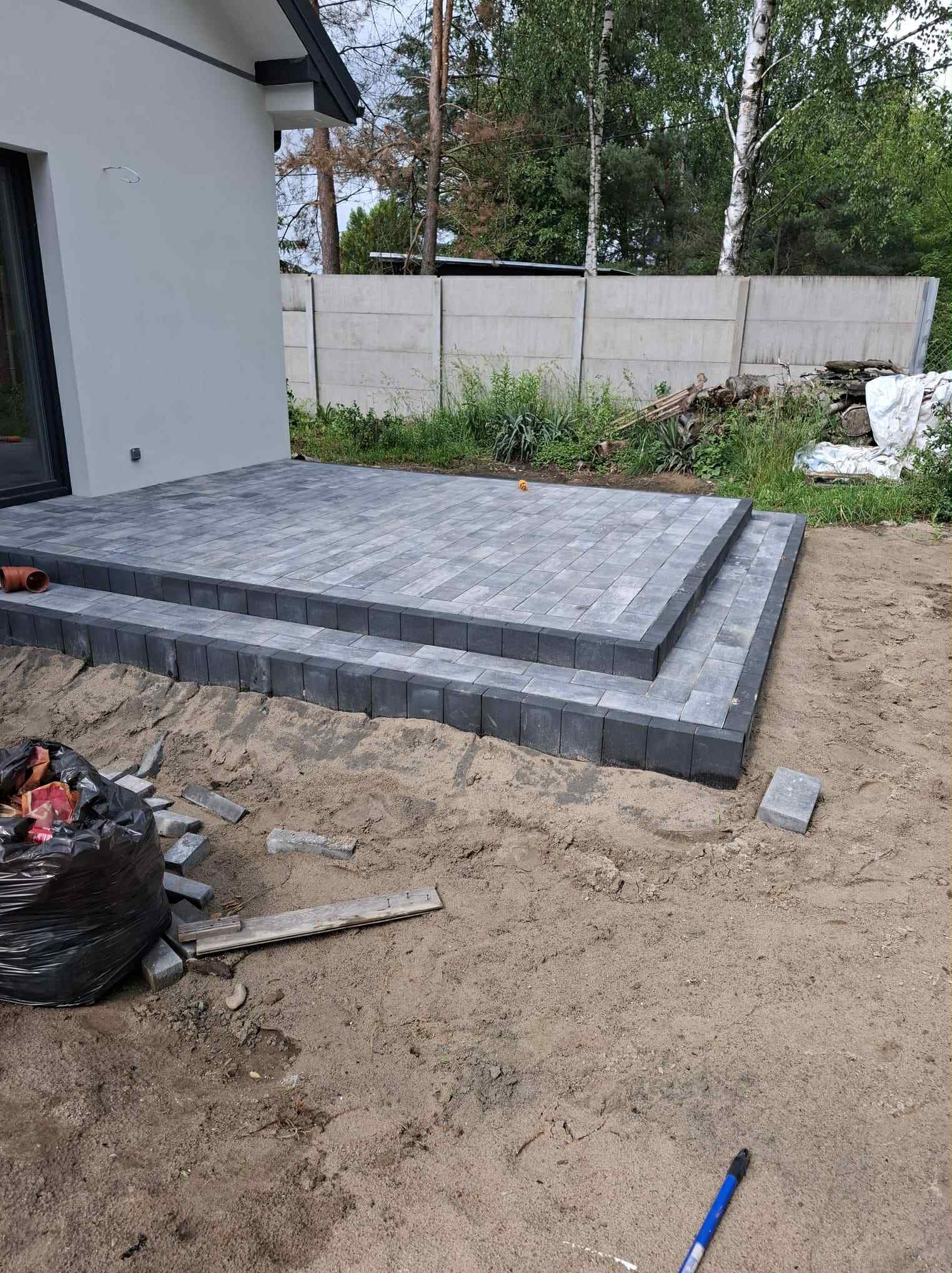 Schody z szarej kostki brukowej prowadzące do domu, widoczne narzędzia i materiały budowlane. Tło stanowi ogrodzenie i zieleń.