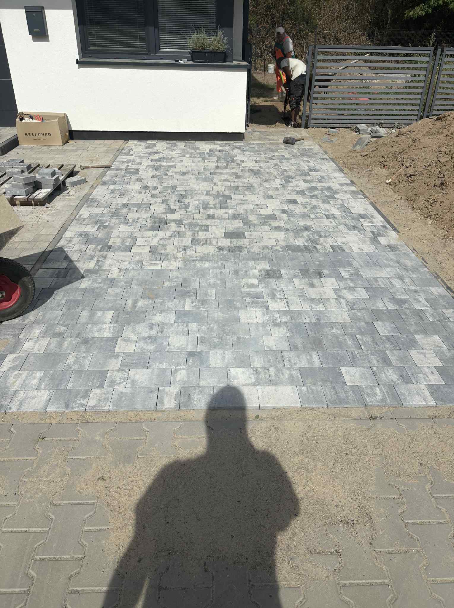 Ułożona szara kostka brukowa na podjeździe domu, widoczni pracownicy w oddali i sterta kostki obok budynku. Cień osoby w dolnej części kadru.