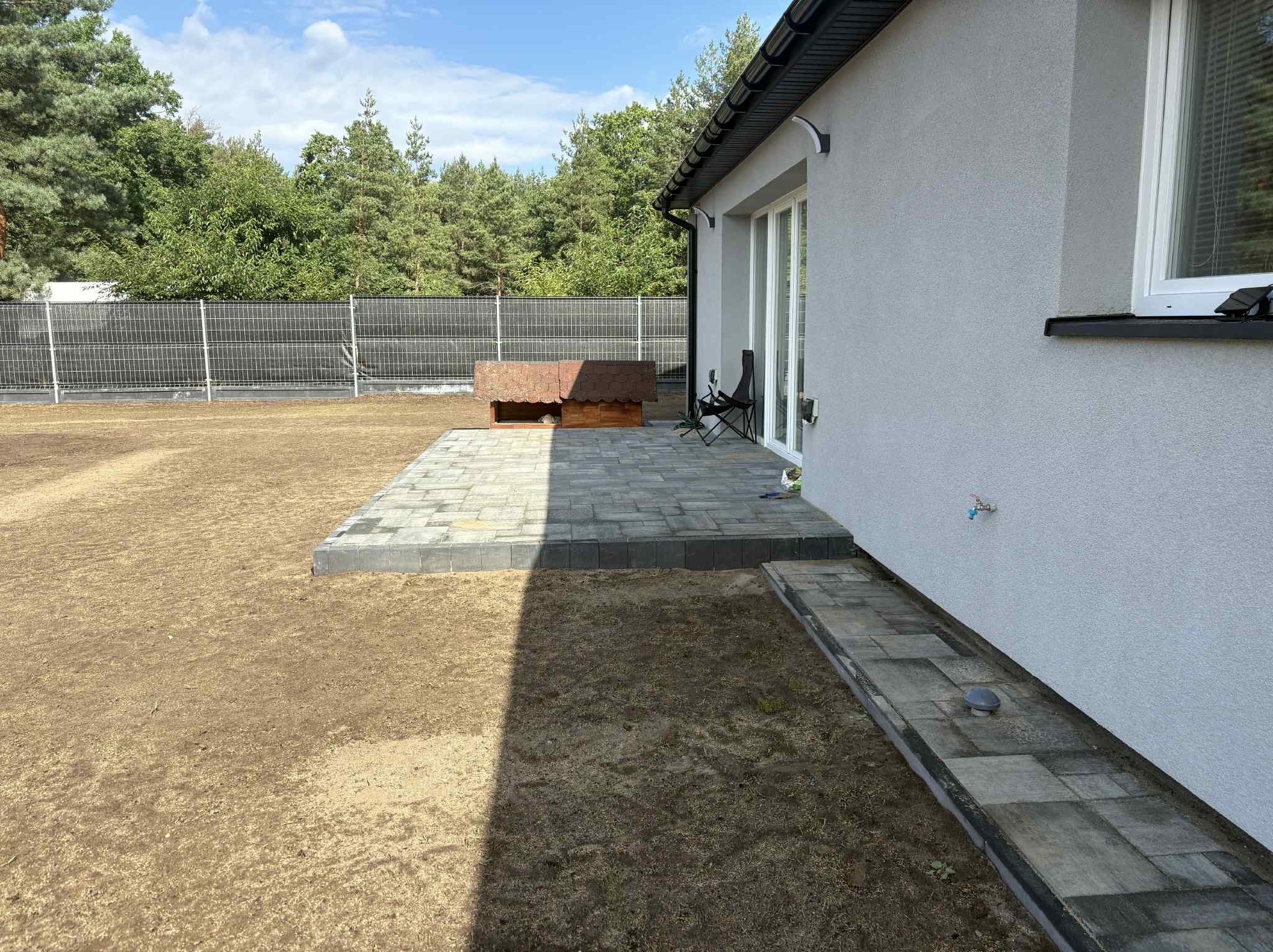 Ułożona kostka brukowa wzdłuż domu z szarą elewacją i brązową budą dla psa. Widoczny trawnik i ogrodzenie z siatki.