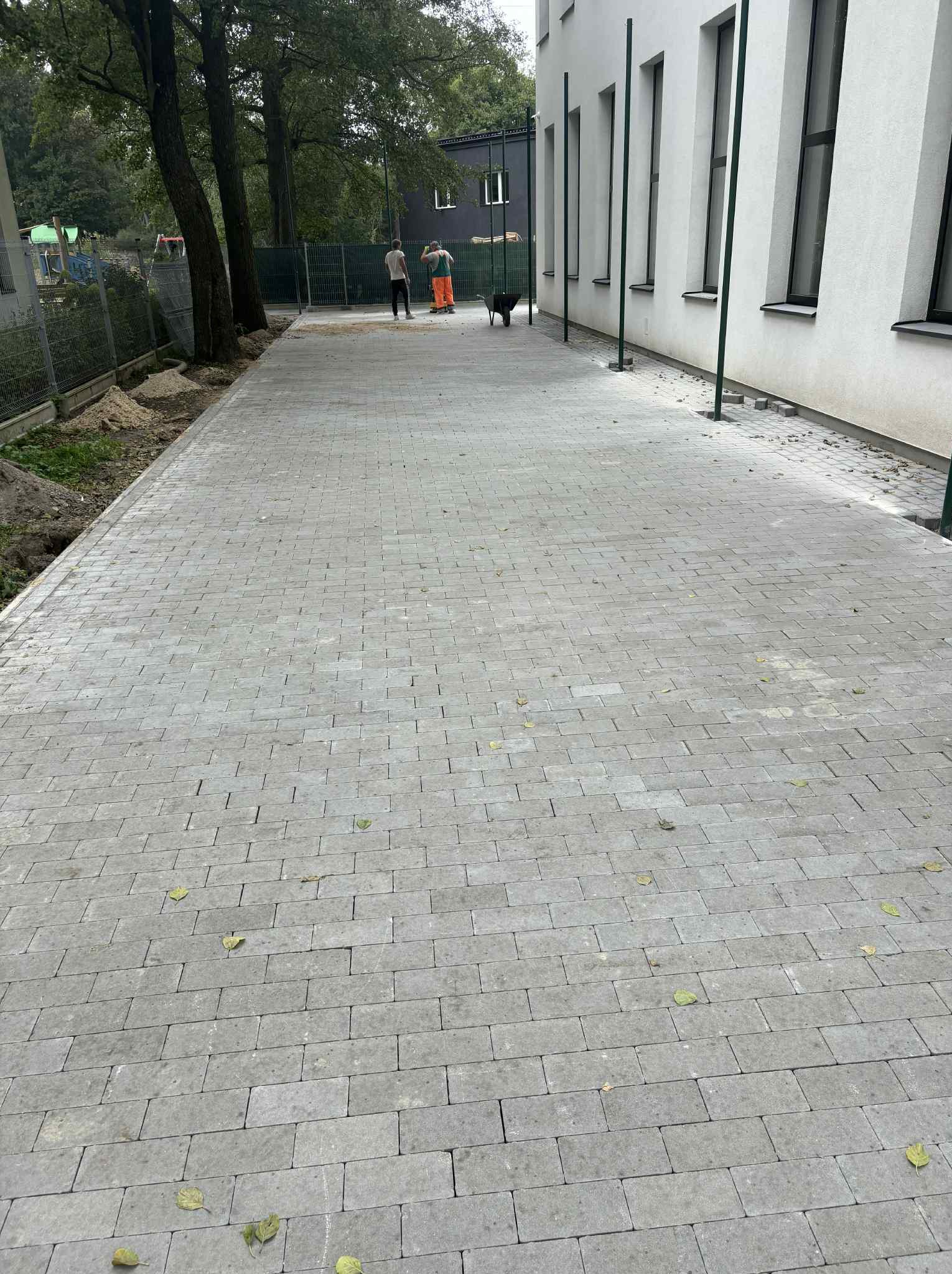 Ułożona szara kostka brukowa na długim chodniku przy budynku. W tle pracownicy i taczka. Widoczne opadłe liście na kostce.