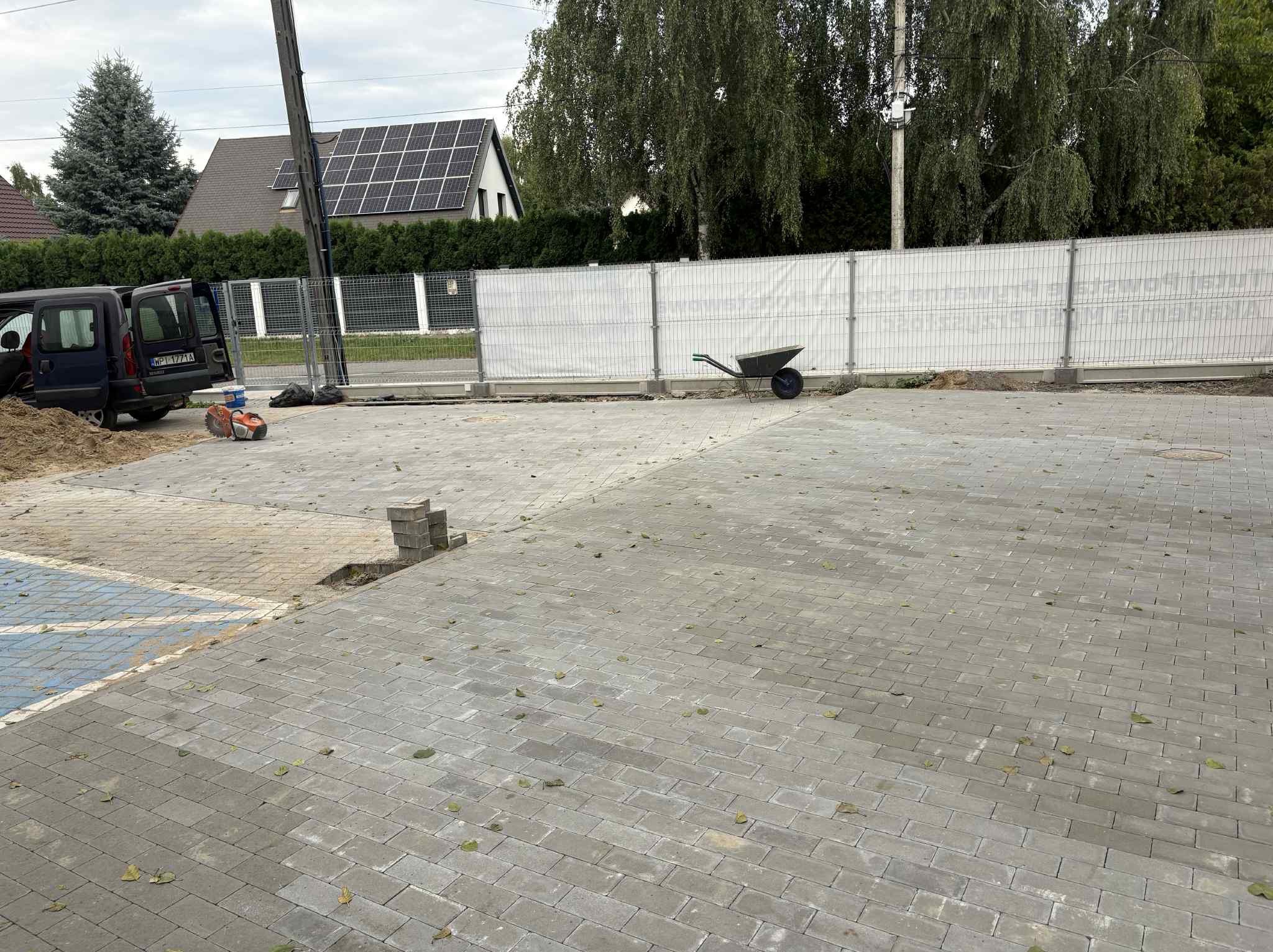 Ułożona szara kostka brukowa na podjeździe, z widocznymi narzędziami i samochodem w tle. Częściowo ułożona kostka przy miejscu parkingowym dla niepełnosprawnych.