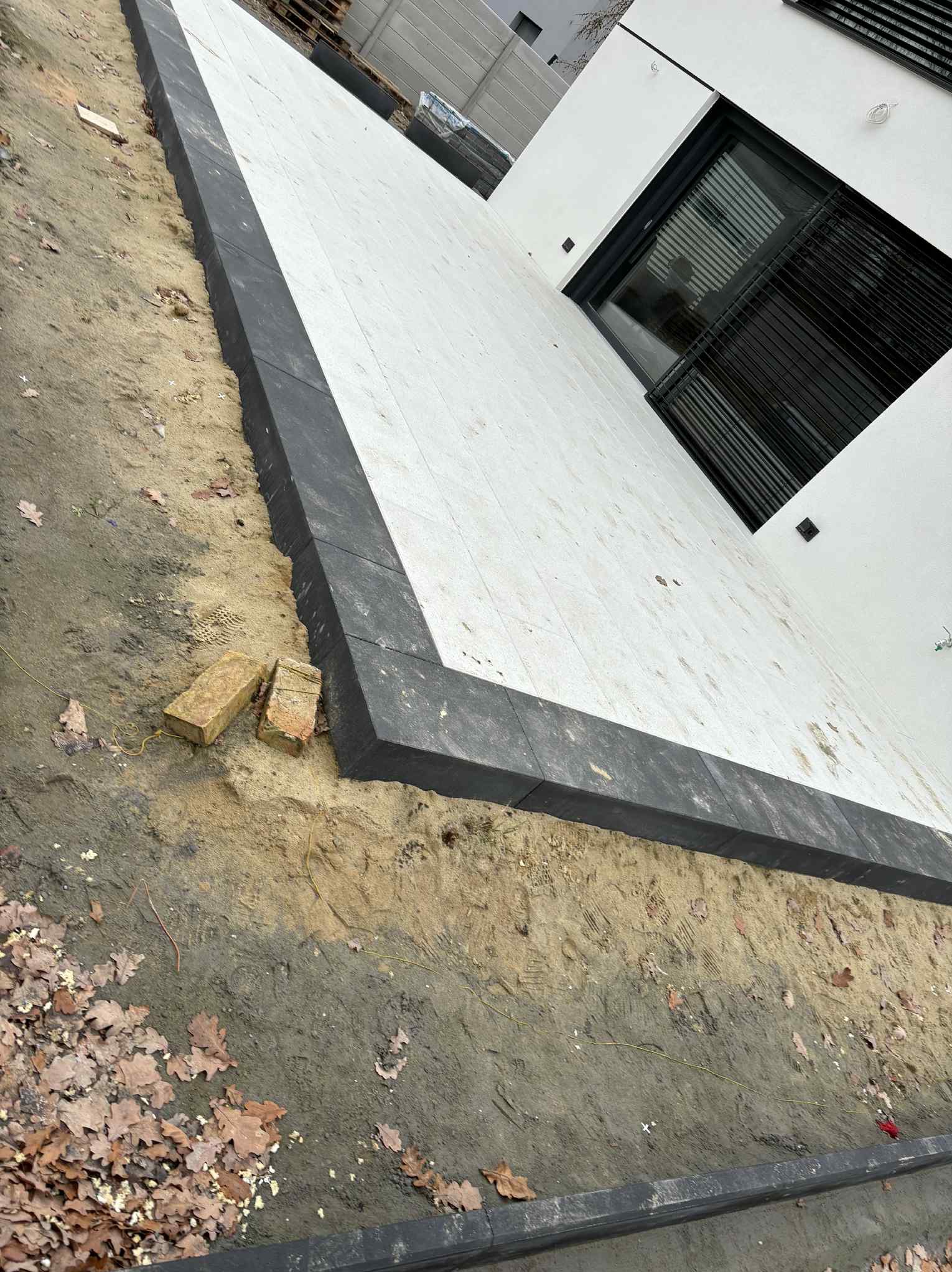 Nowoczesny taras z jasnych płyt obramowany ciemną kostką, widok z góry. Częściowo ułożona nawierzchnia, widoczne ślady prac ziemnych i cegły. Minimalistyczny design.