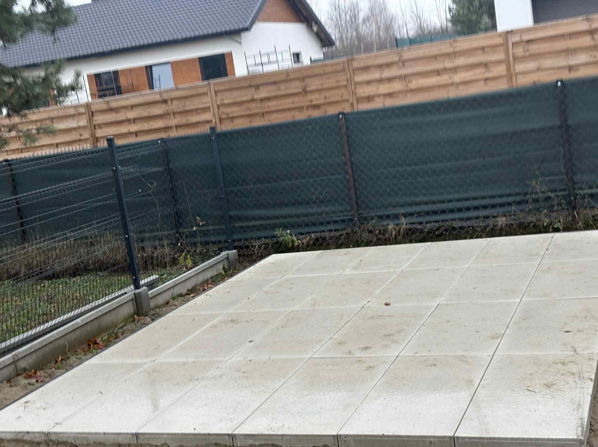 Świeżo ułożona, jasna kostka brukowa na posesji, widok z góry. W tle ogrodzenie z siatki i drewniane panele, dom z ciemnym dachem. Pochmurny dzień.